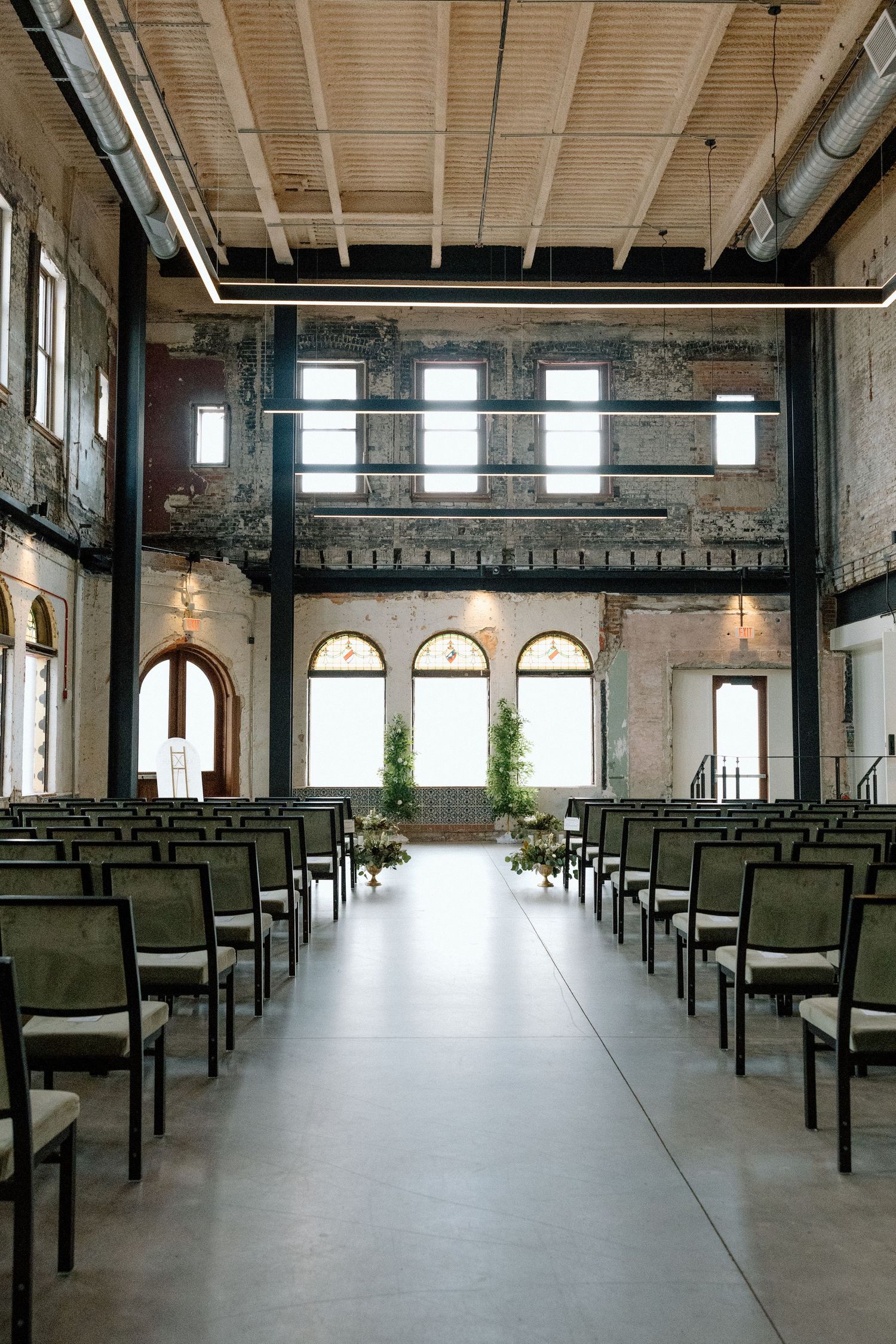 Elegant rustic wedding ceremony venue with rows of green chairs, exposed brick walls, and arched windows.