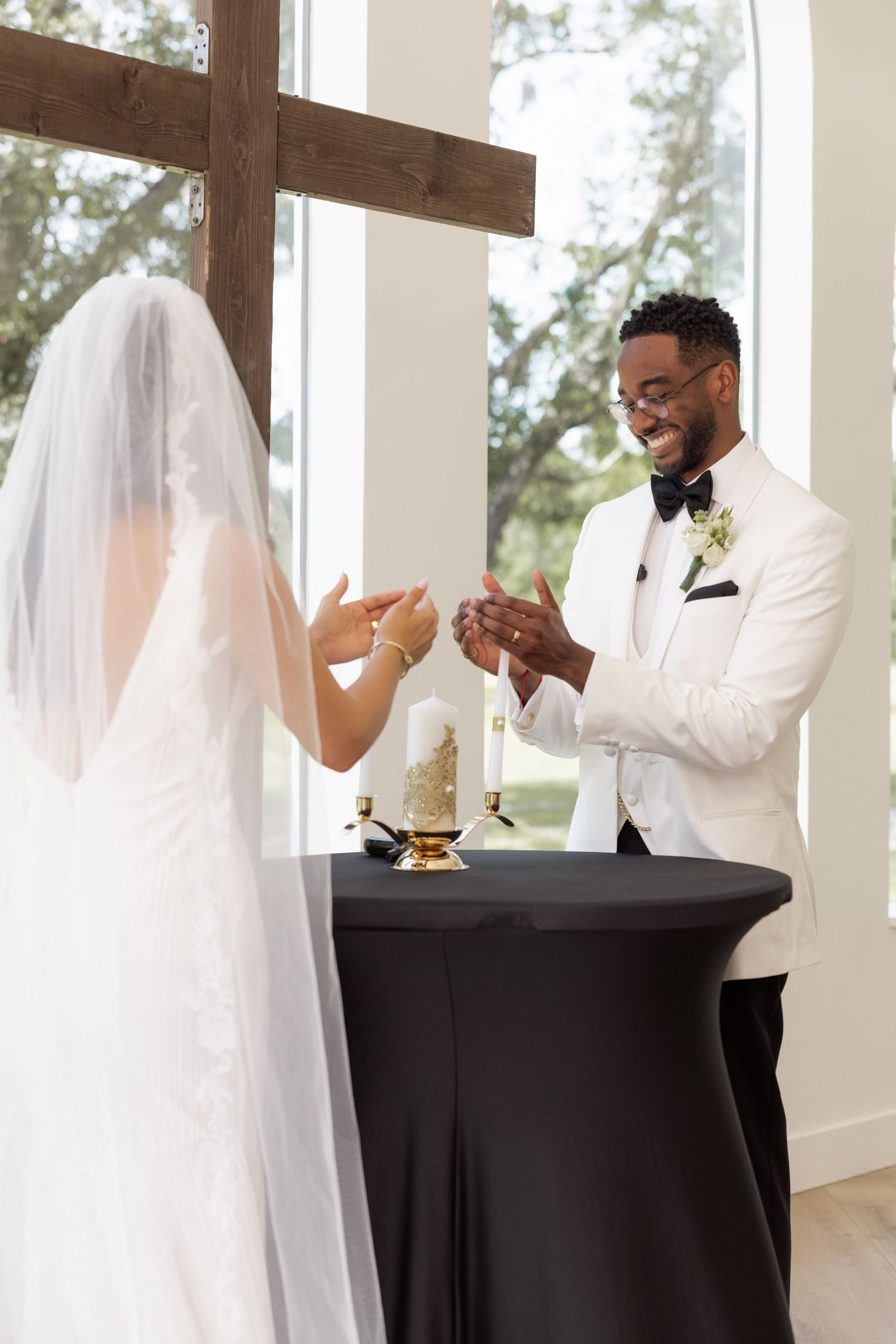 Groom in white tuxedo smiles at bride during unity candle ceremony in bright chapel.