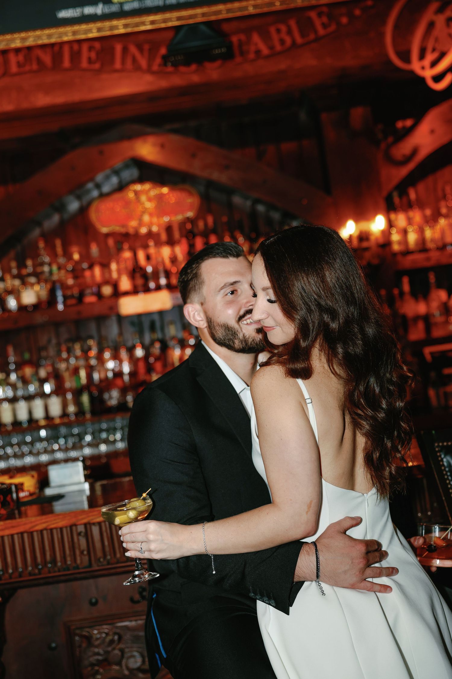 Couple embraces intimately at a candlelit bar, man holding a cocktail while woman wraps arms around him.