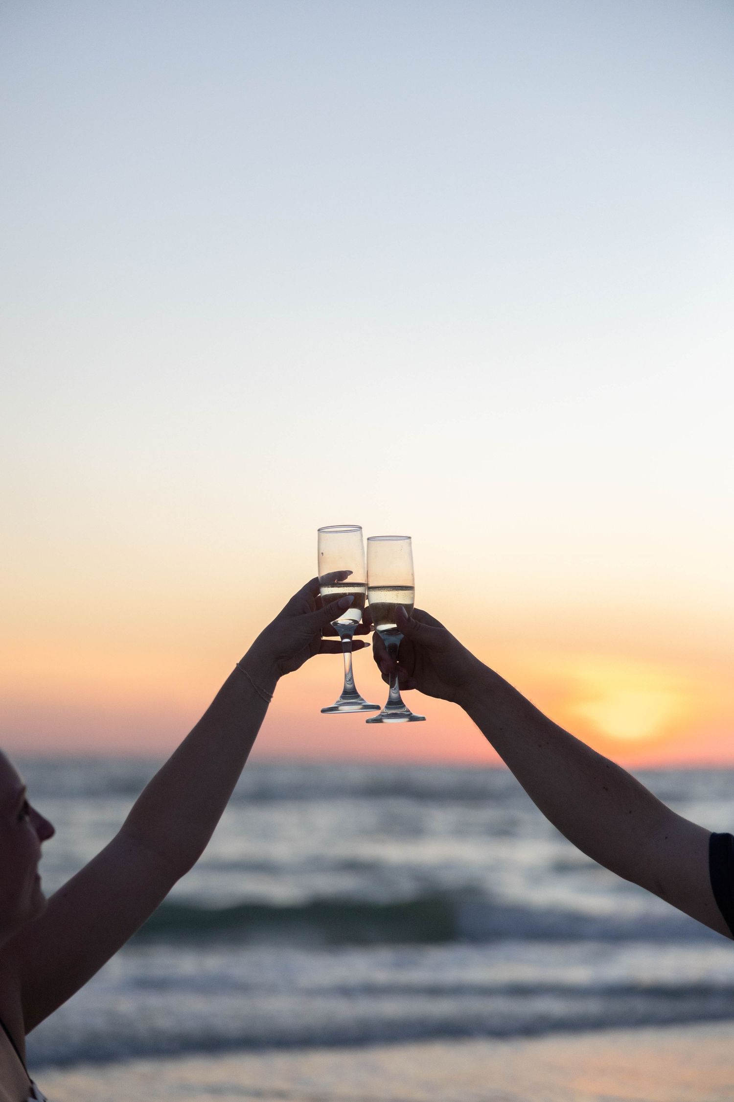 Two hands clinking champagne glasses together against a stunning golden sunset over the ocean beach.