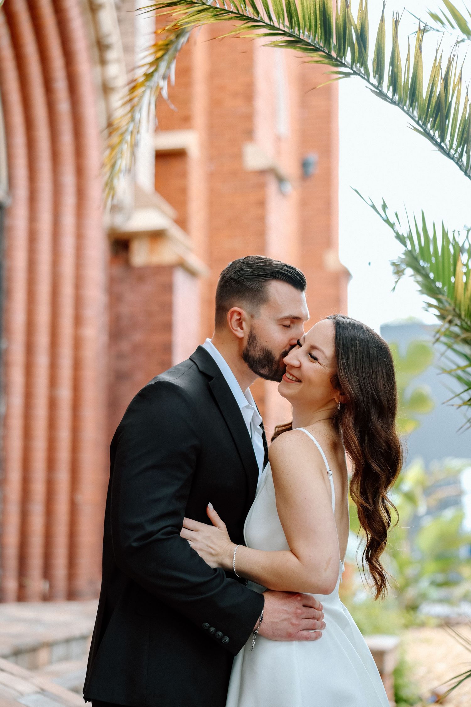 Bride and groom embrace romantically near a red brick building with palm trees at their engagement shoot.