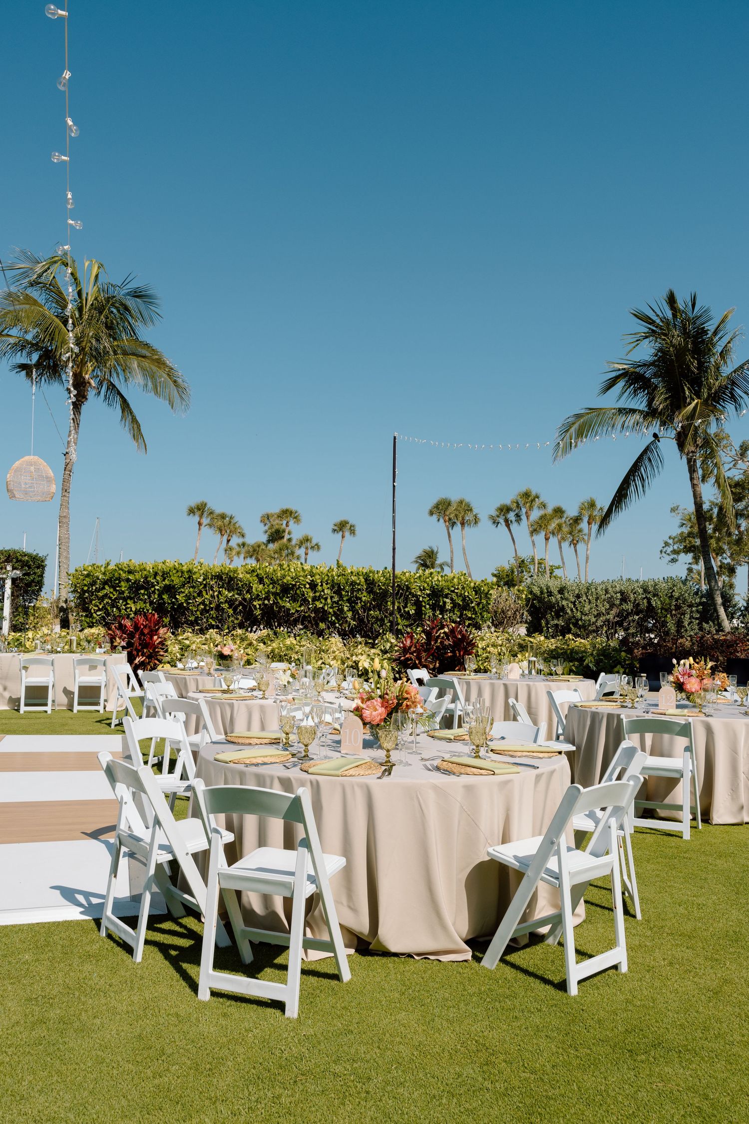 Elegant outdoor wedding reception setup with white chairs, neutral linens, tropical floral centerpieces, and palm trees under blue sky.