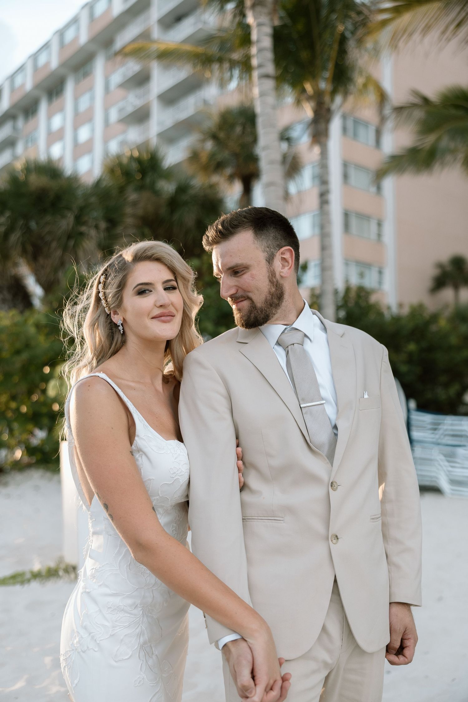 Elegant bride and bearded groom in tan suit holding hands on beach with palm trees and resort hotel backdrop.
