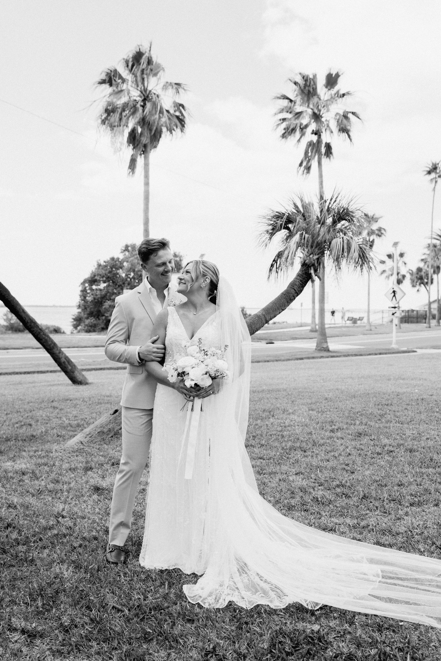 Black and white portrait of bride and groom embracing under tall palm trees on wedding day.