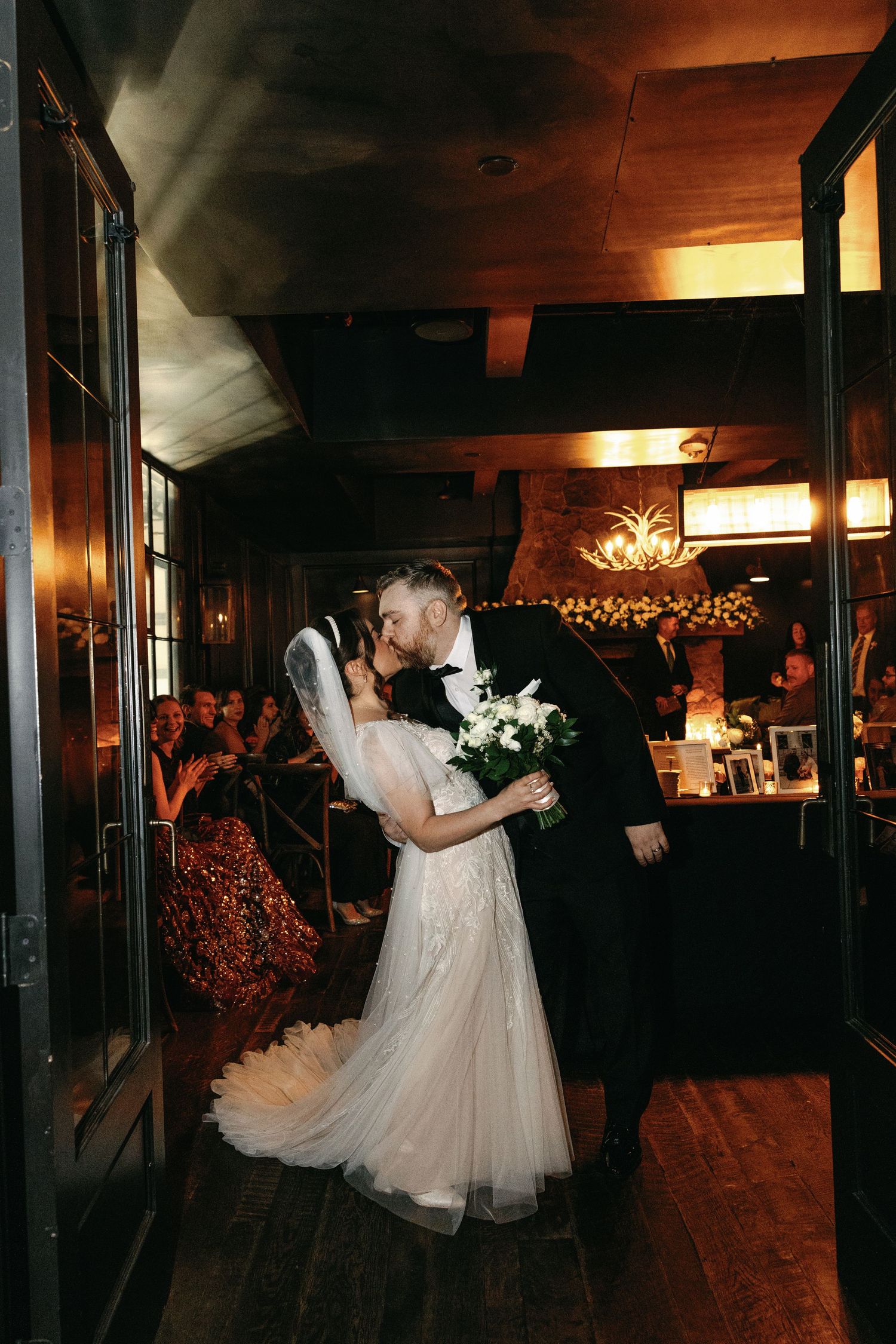 Bride and groom share a kiss at the entrance of a warmly lit rustic wedding reception venue.