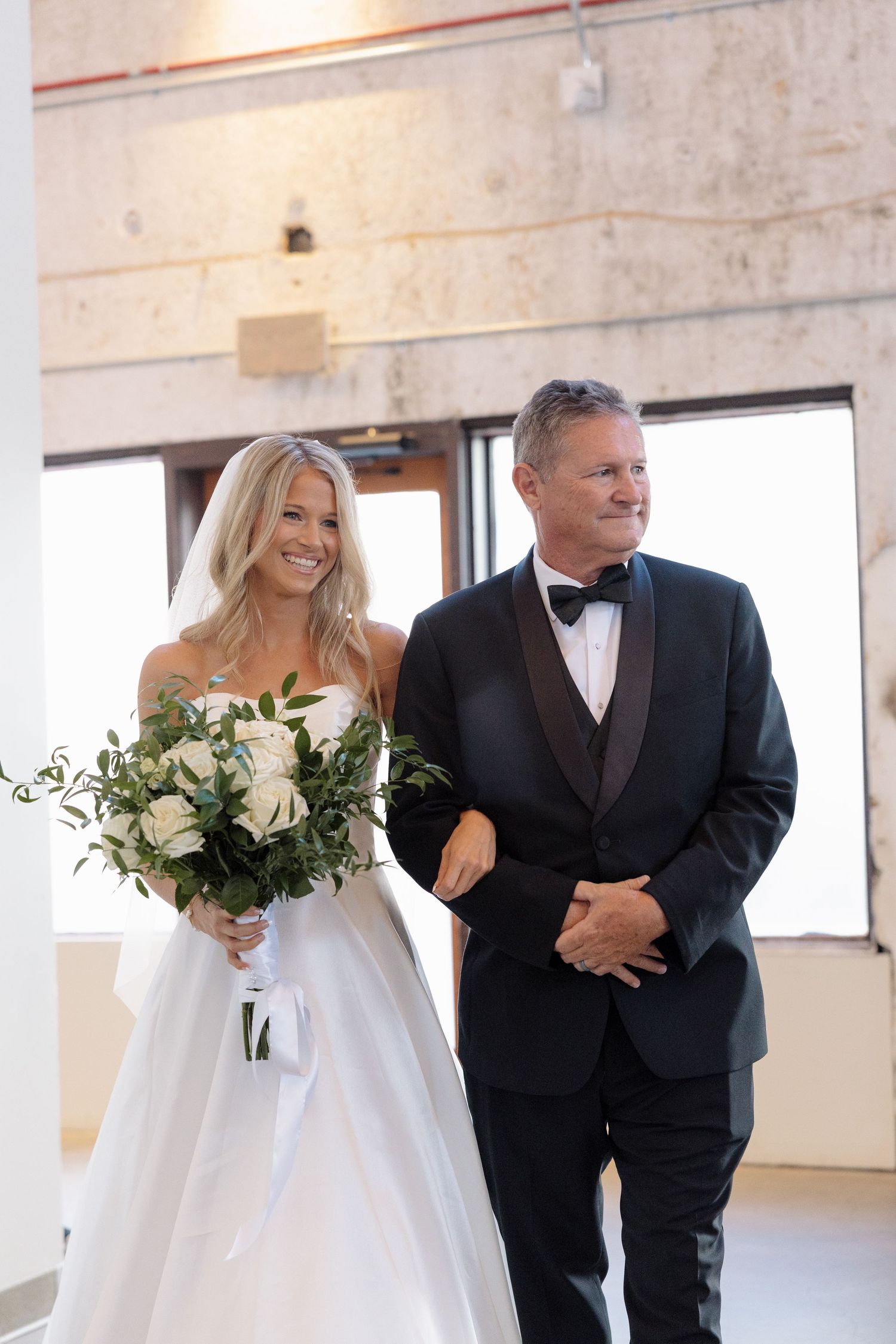 Smiling bride in strapless white gown walks down aisle with father in black tuxedo holding white bouquet.