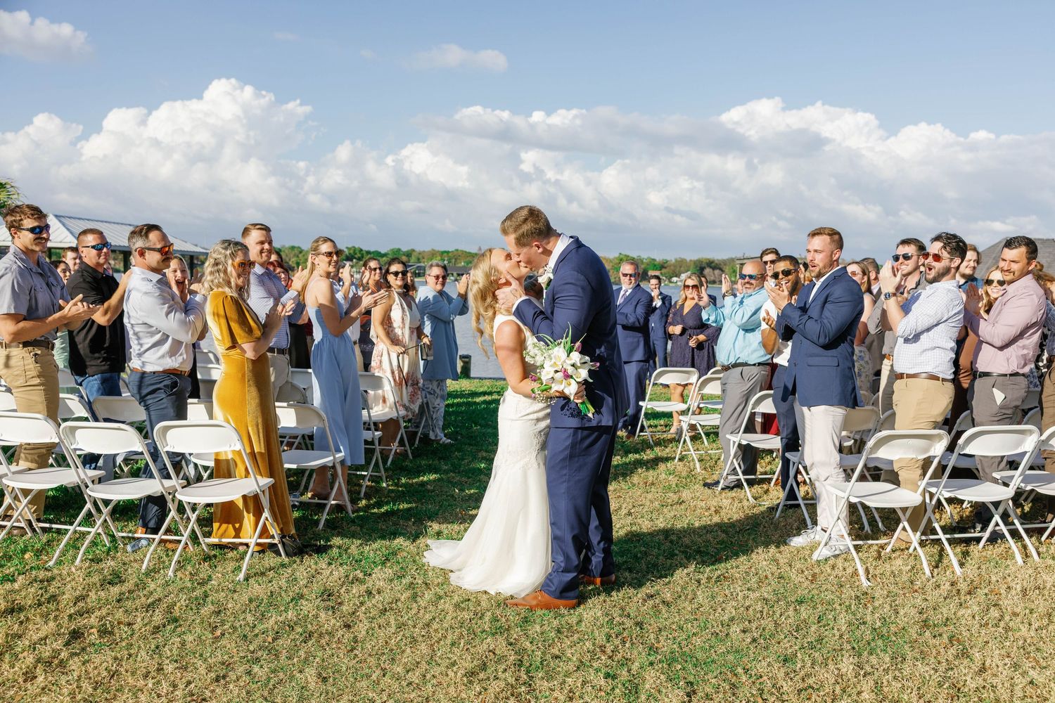 Bride and groom share a kiss mid-aisle surrounded by cheering guests at outdoor lakeside wedding ceremony.
