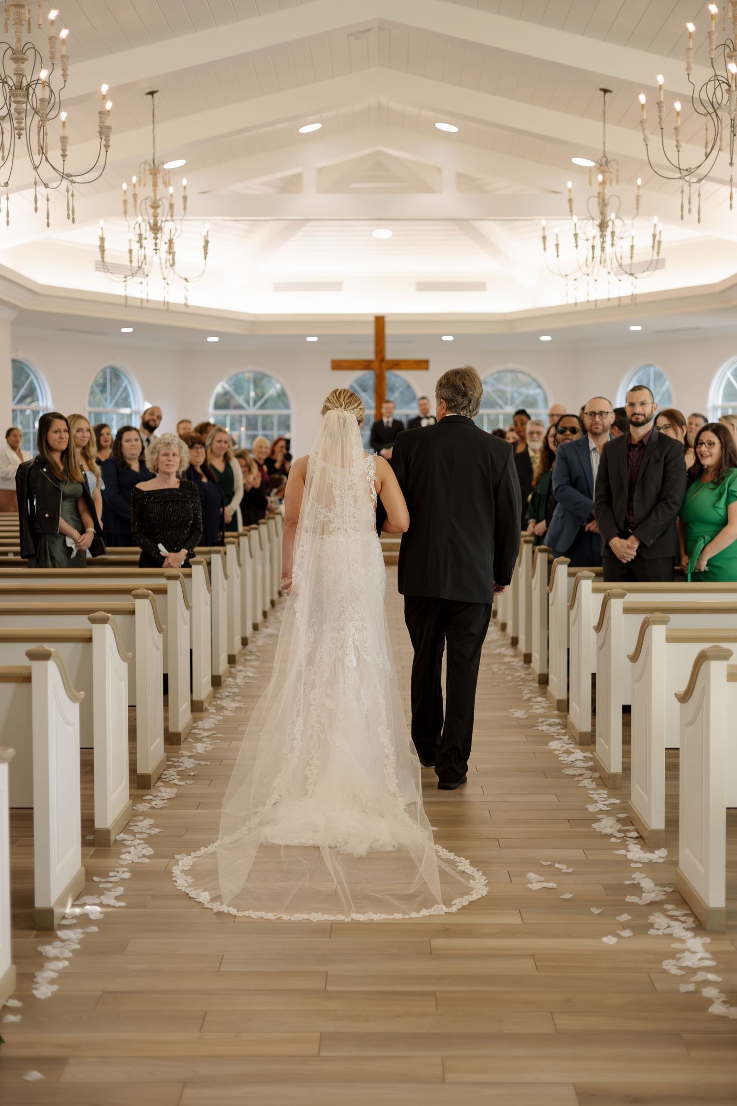 A bride in a lace gown and cathedral veil walks down the aisle with her father.