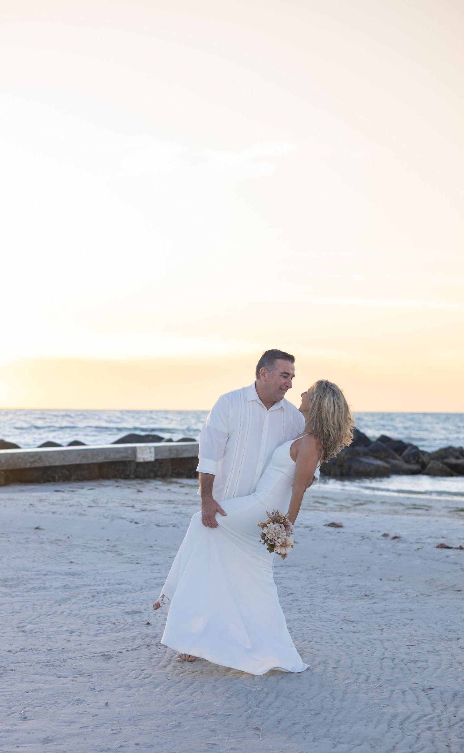 Wedding couple in white attire embracing on beach at sunset with rocky jetty in background.