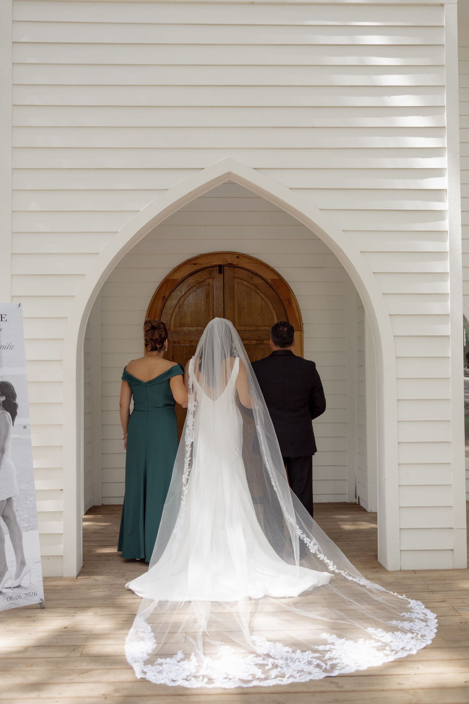Bride in lace veil with parents entering arched doorway of white chapel before ceremony.