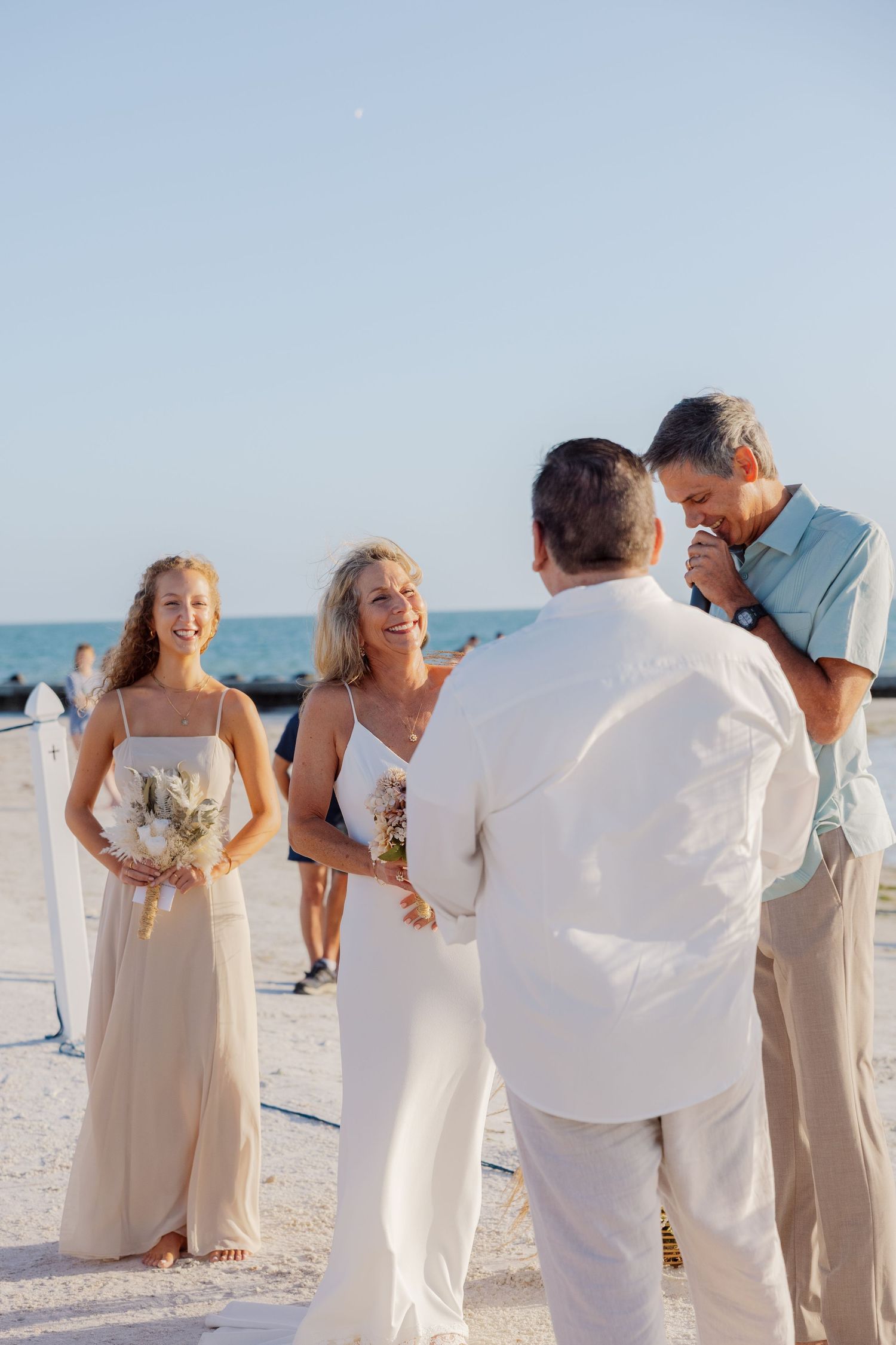 Bride laughs joyfully during emotional beach wedding ceremony with bridesmaid and officiant.