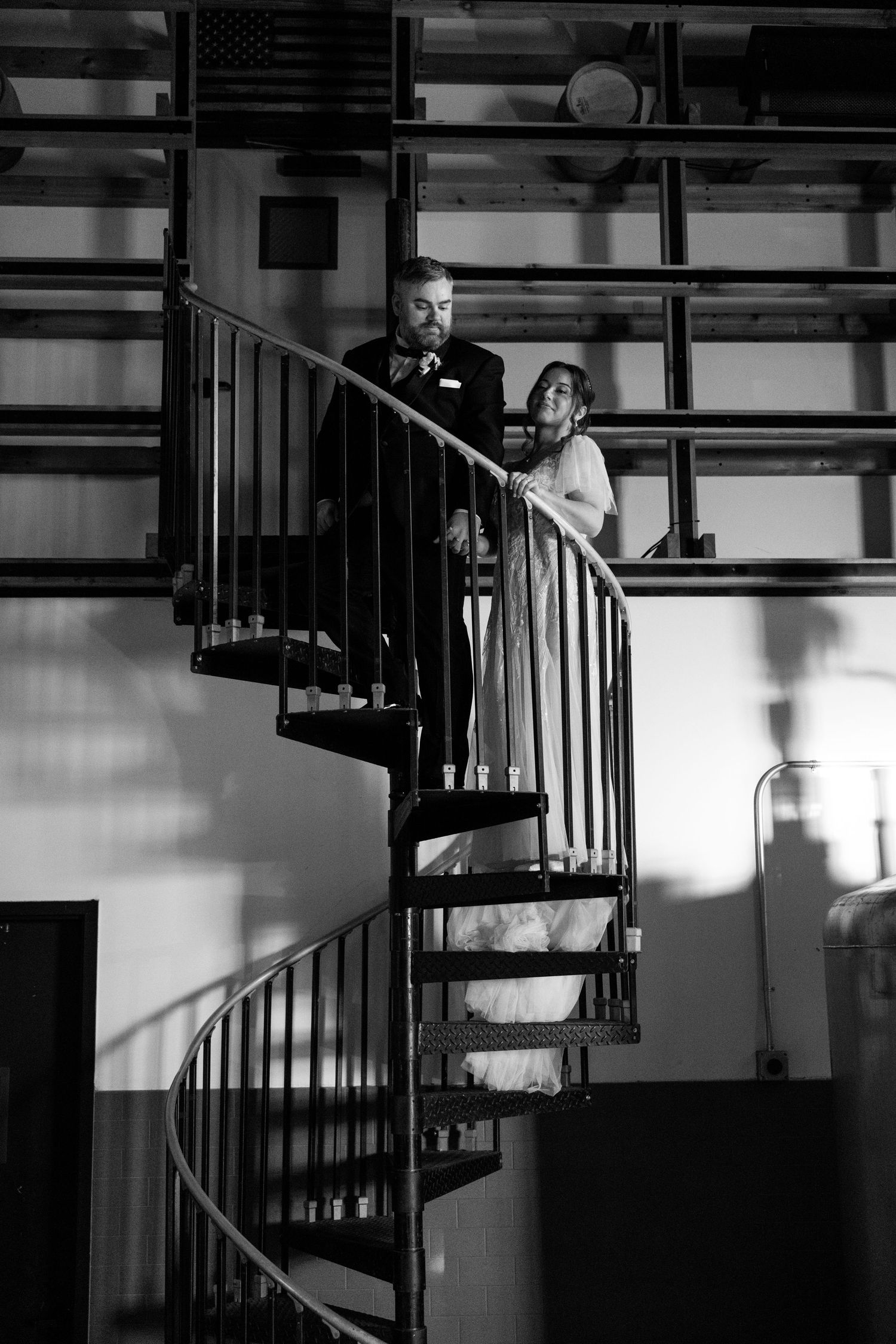 Black and white photo of bride and groom standing together on a spiral staircase inside an industrial venue.