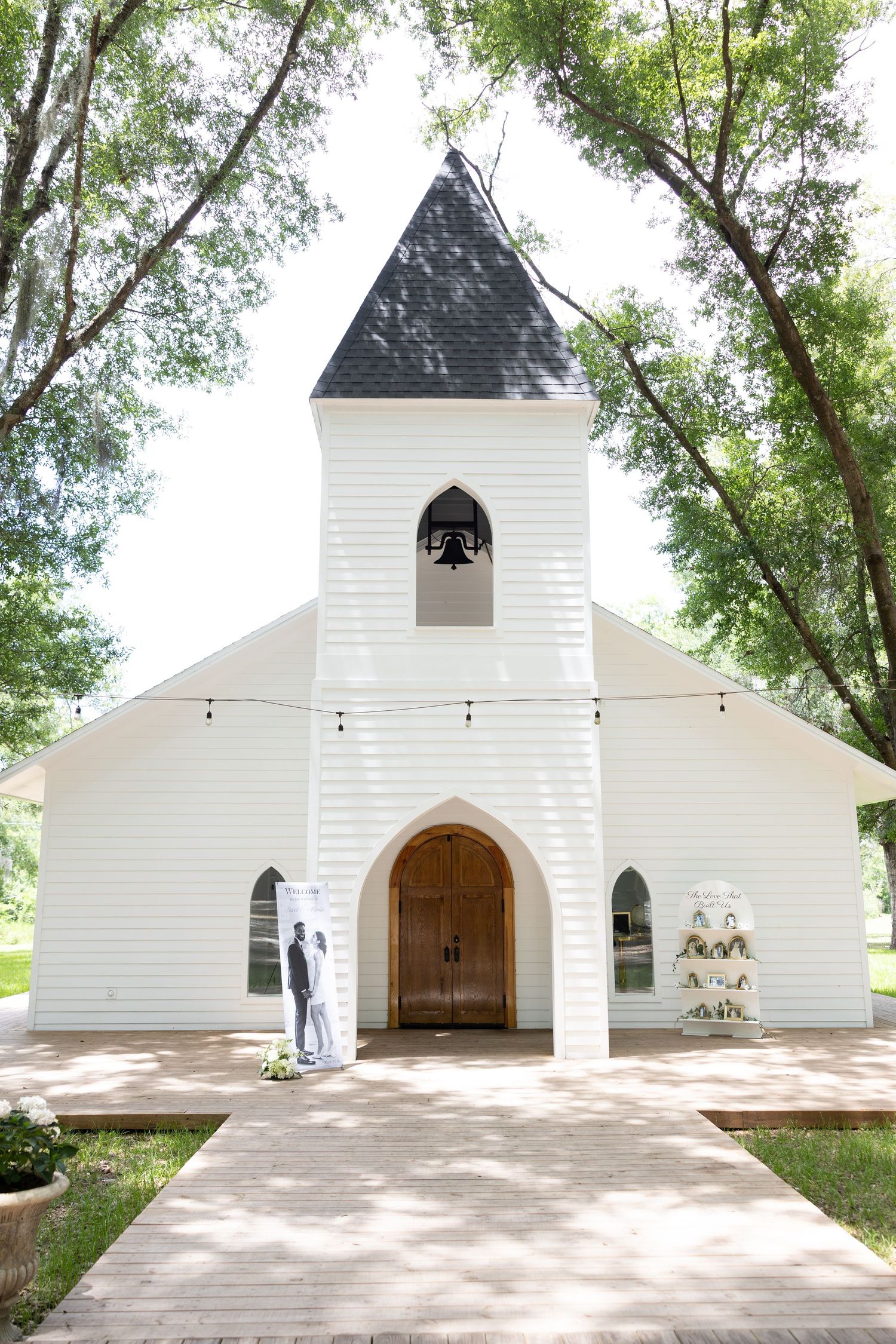 White wedding chapel with dark spire, string lights, and wooden doors framed by tall trees.