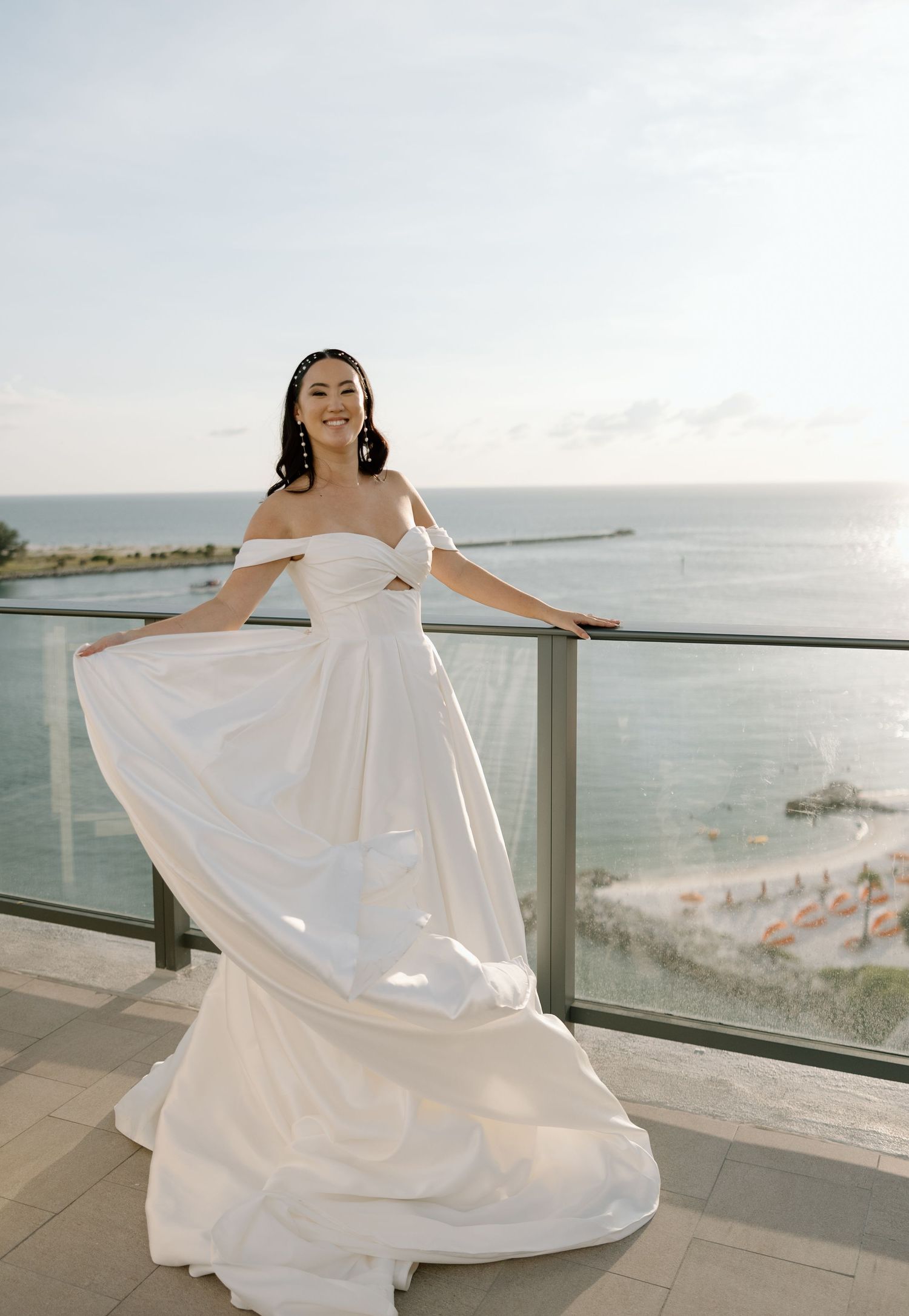 Bride in flowing off-shoulder white gown twirls on balcony with ocean and beach visible behind her.