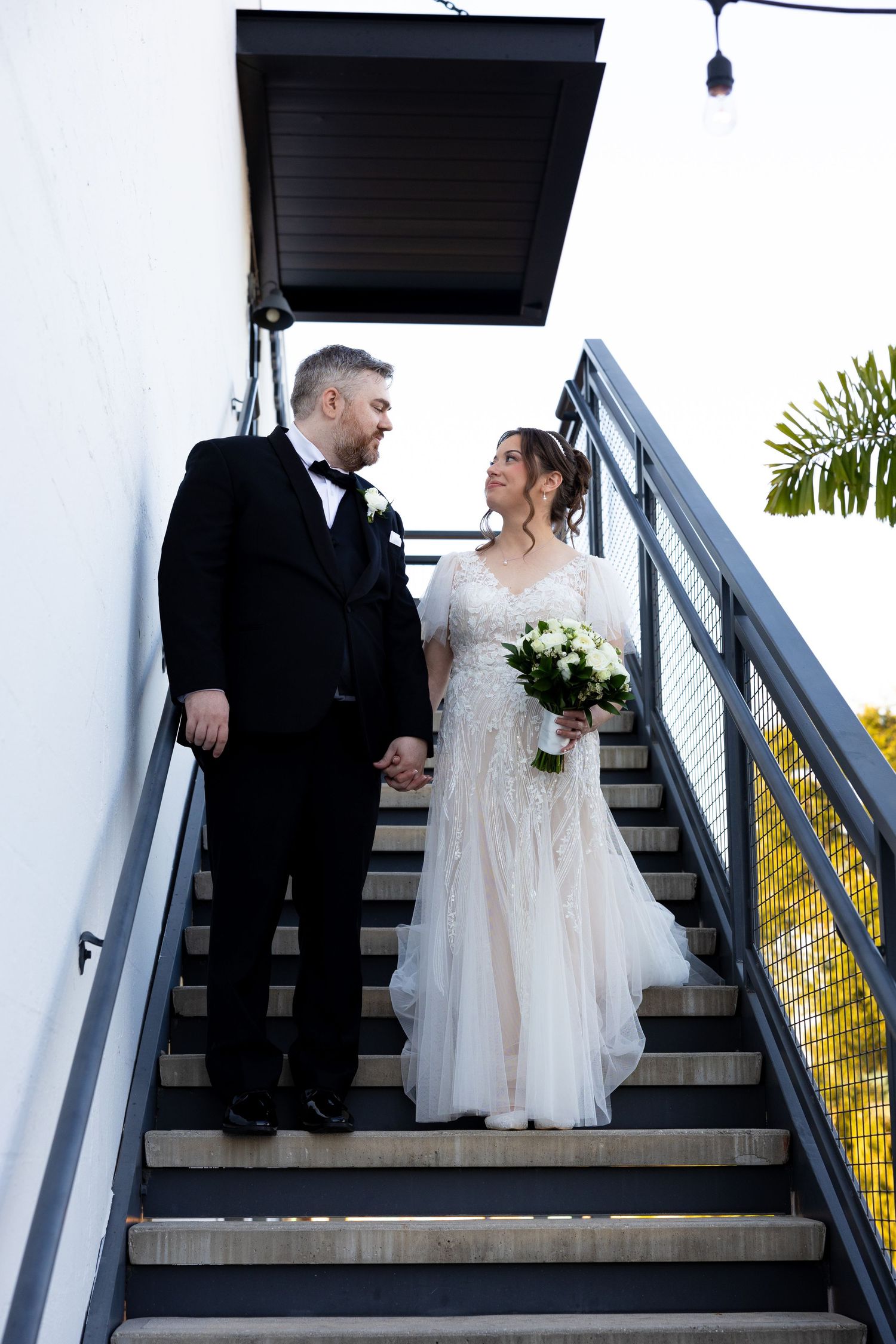 Bride and groom hold hands on outdoor staircase, she in lace gown with bouquet, he in black tuxedo.