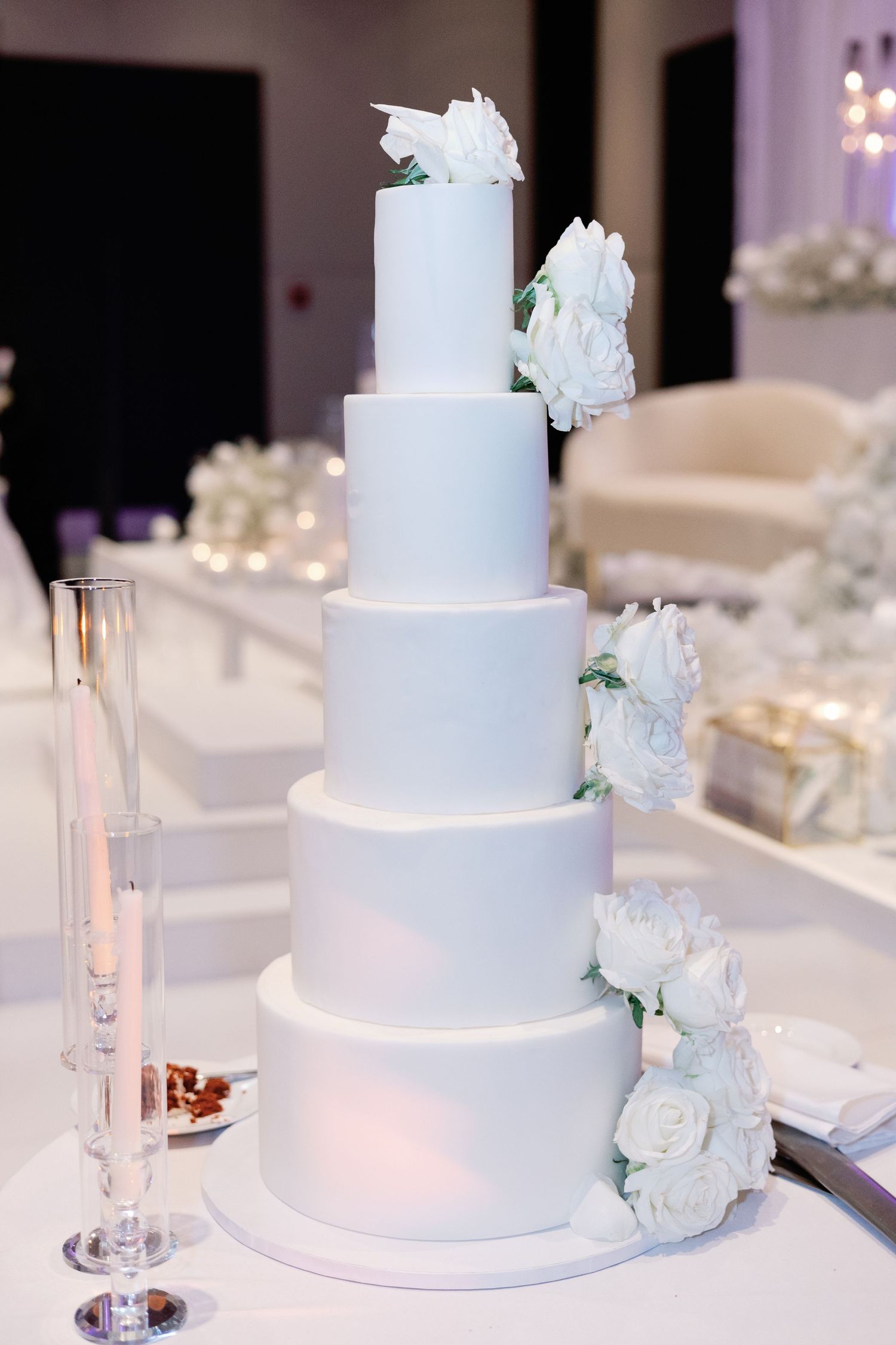 Elegant five-tier white wedding cake adorned with white peony flowers on a decorated reception table.