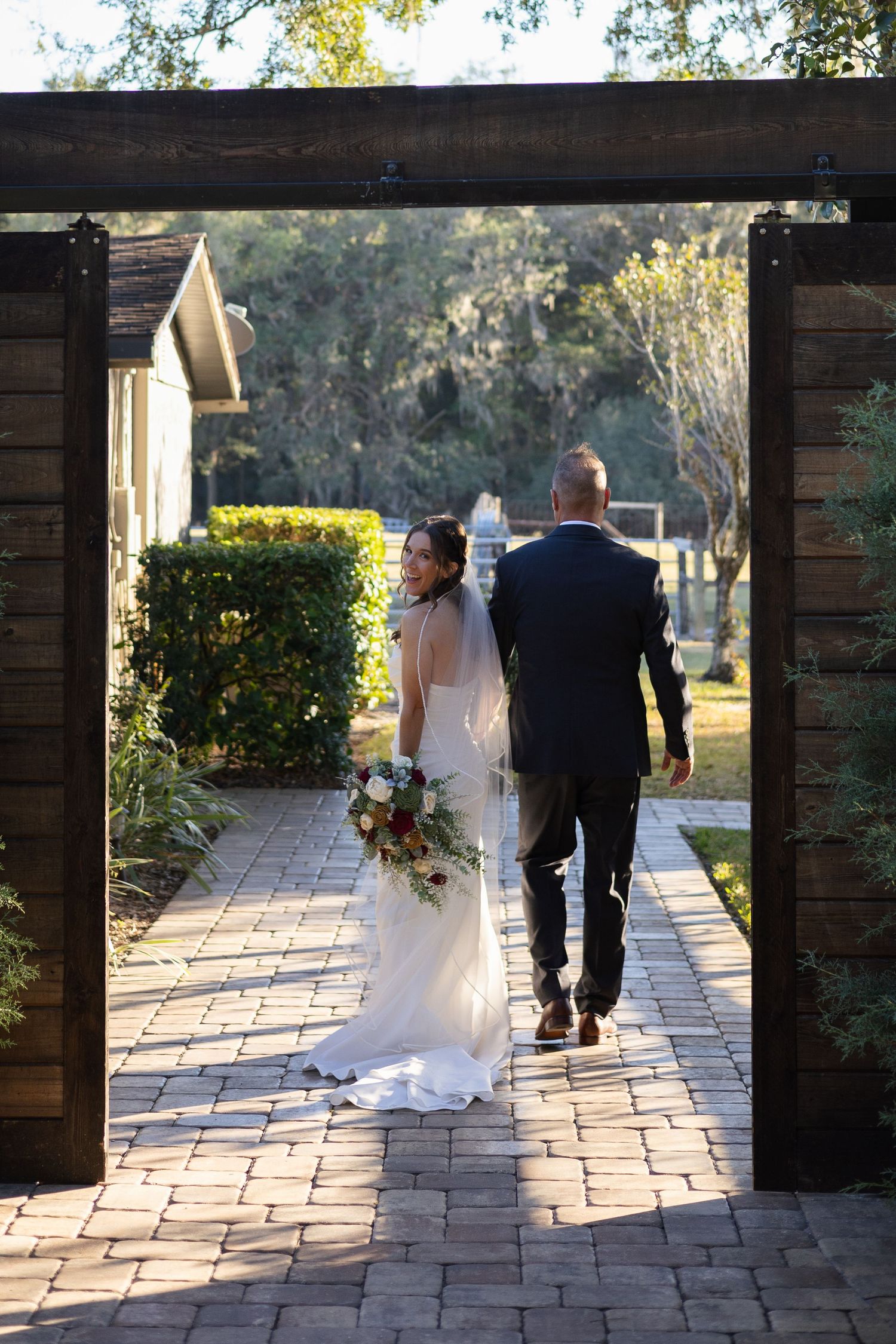 Bride and groom walking hand-in-hand through rustic wooden barn doors into a sunlit garden pathway.