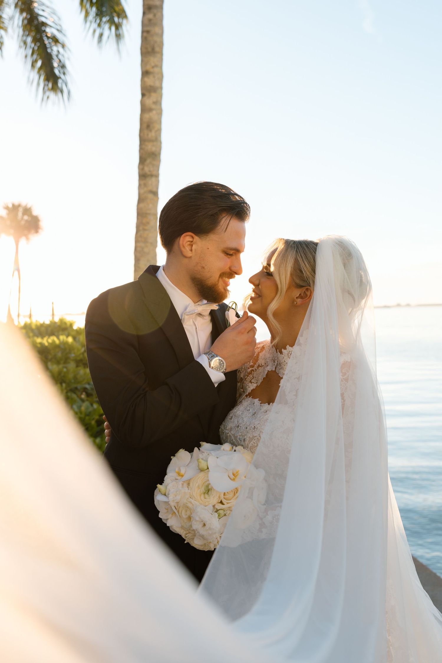 A bride and groom share a romantic moment near the ocean at sunset, with palm trees and golden light in the background.