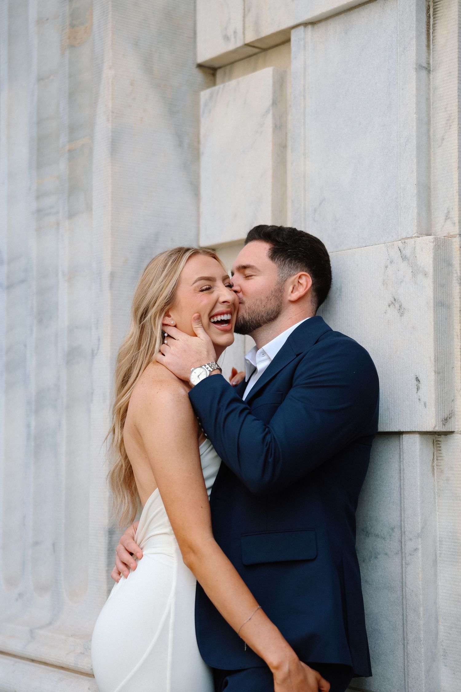 Laughing bride and groom sharing a joyful embrace against a marble wall during engagement photos.