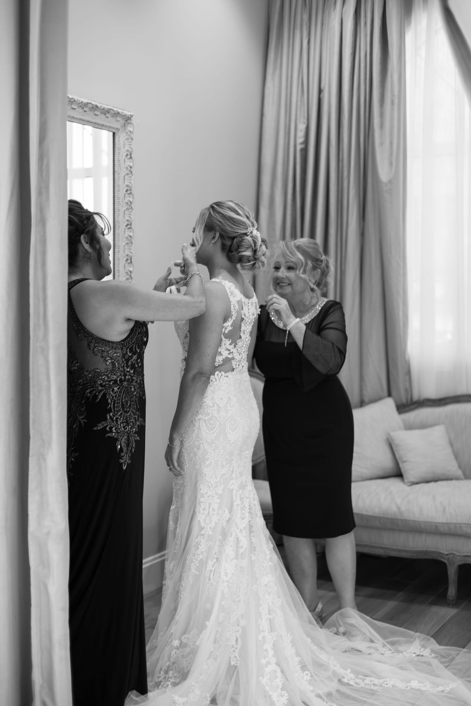 Bride in lace wedding gown being assisted by two women while looking in ornate mirror before ceremony.