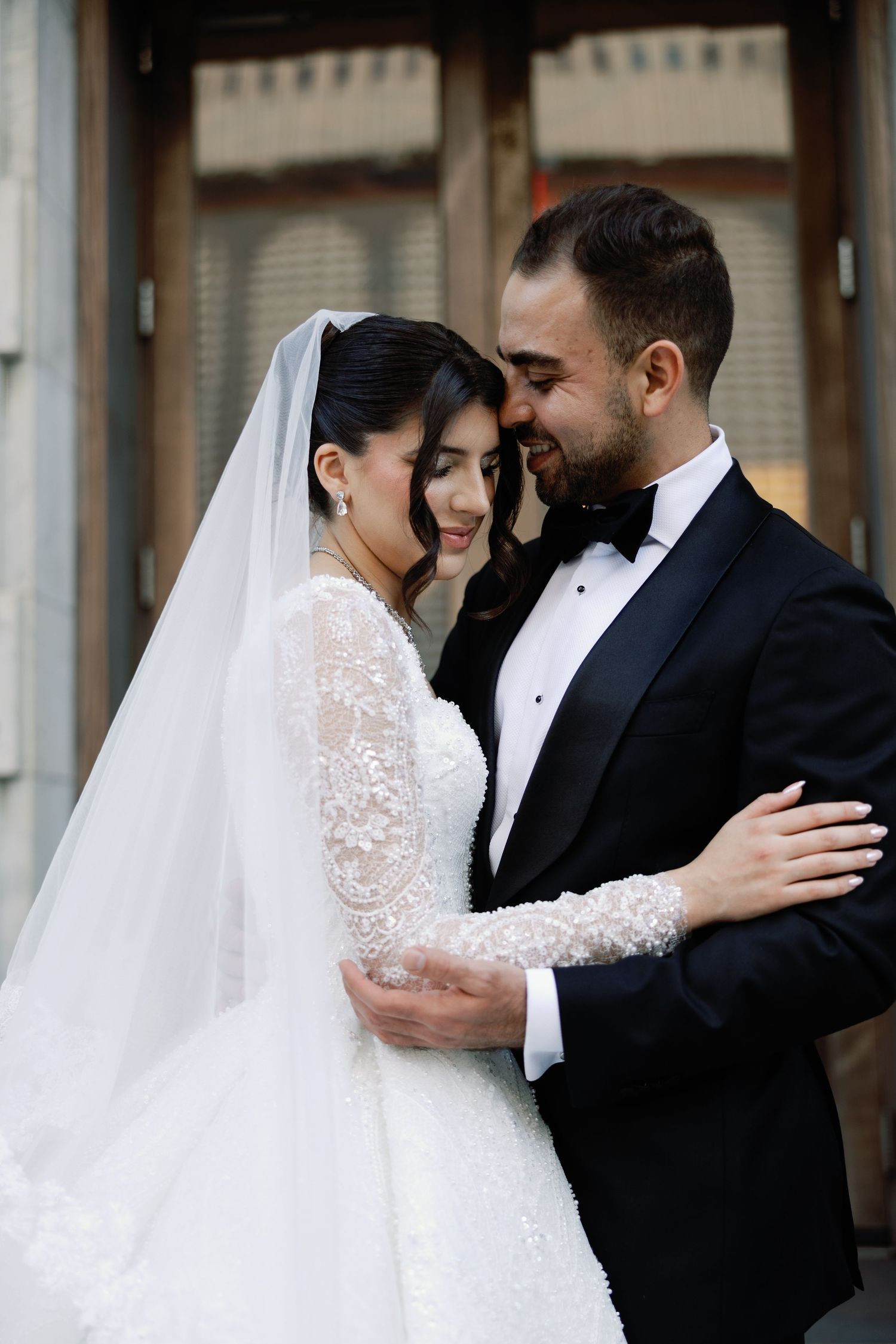 Bride in lace long-sleeve gown and groom in tuxedo sharing a joyful moment outdoors.
