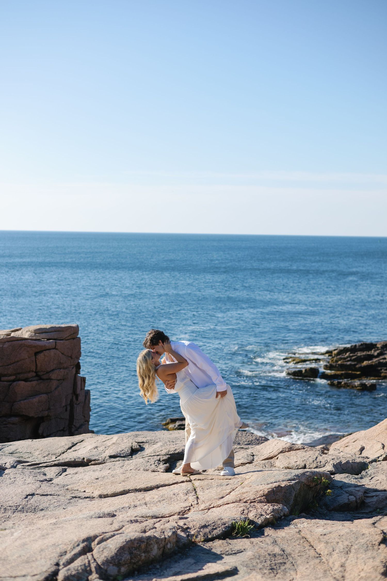 Man dipping woman in white dress for a romantic kiss on rugged seaside rocks by the ocean.