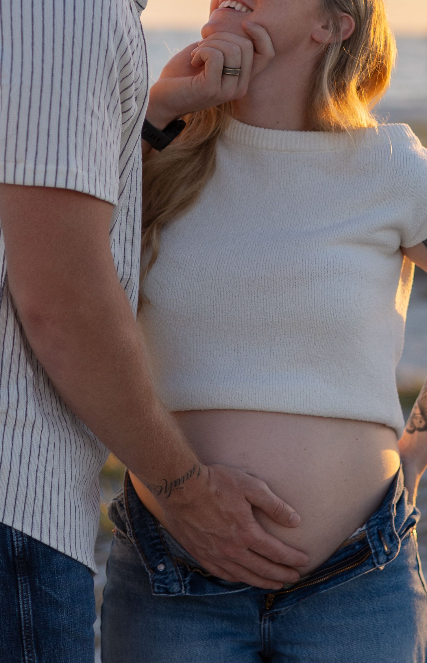 Close-up of pregnant woman's bare belly being tenderly held by partner at golden hour beach.