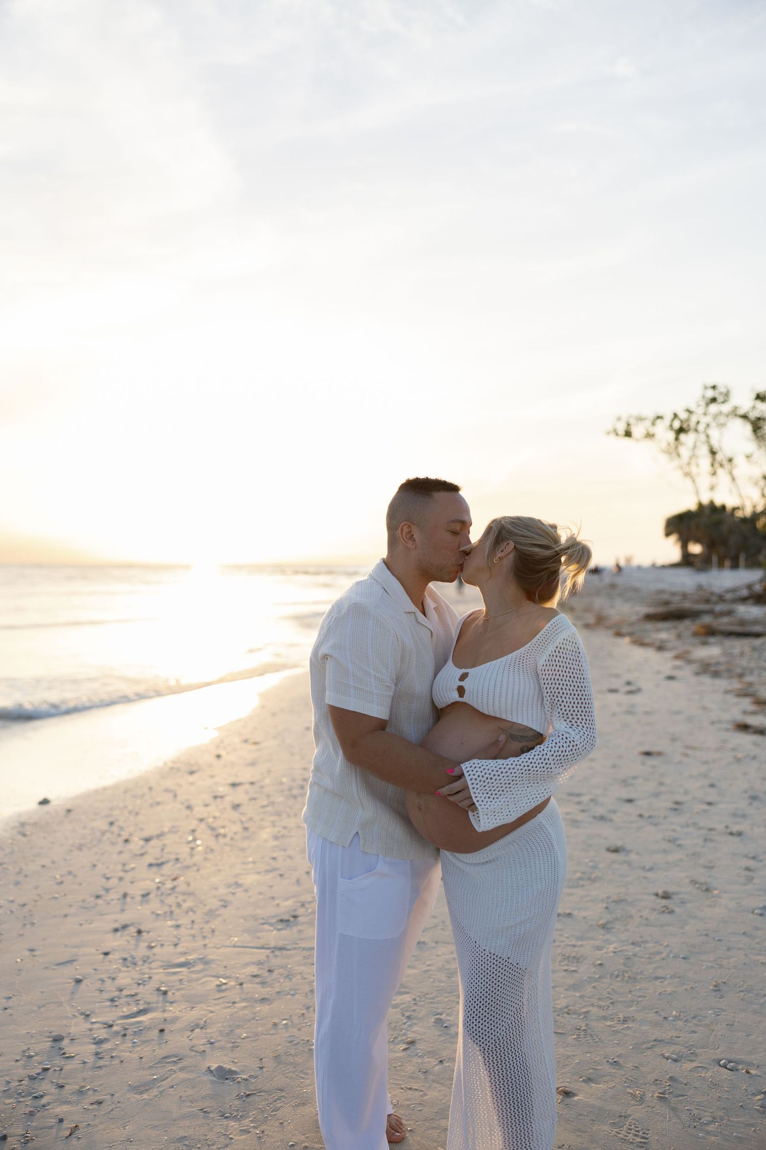 Couple kissing on sandy beach at golden sunset, woman visibly pregnant in white outfit.