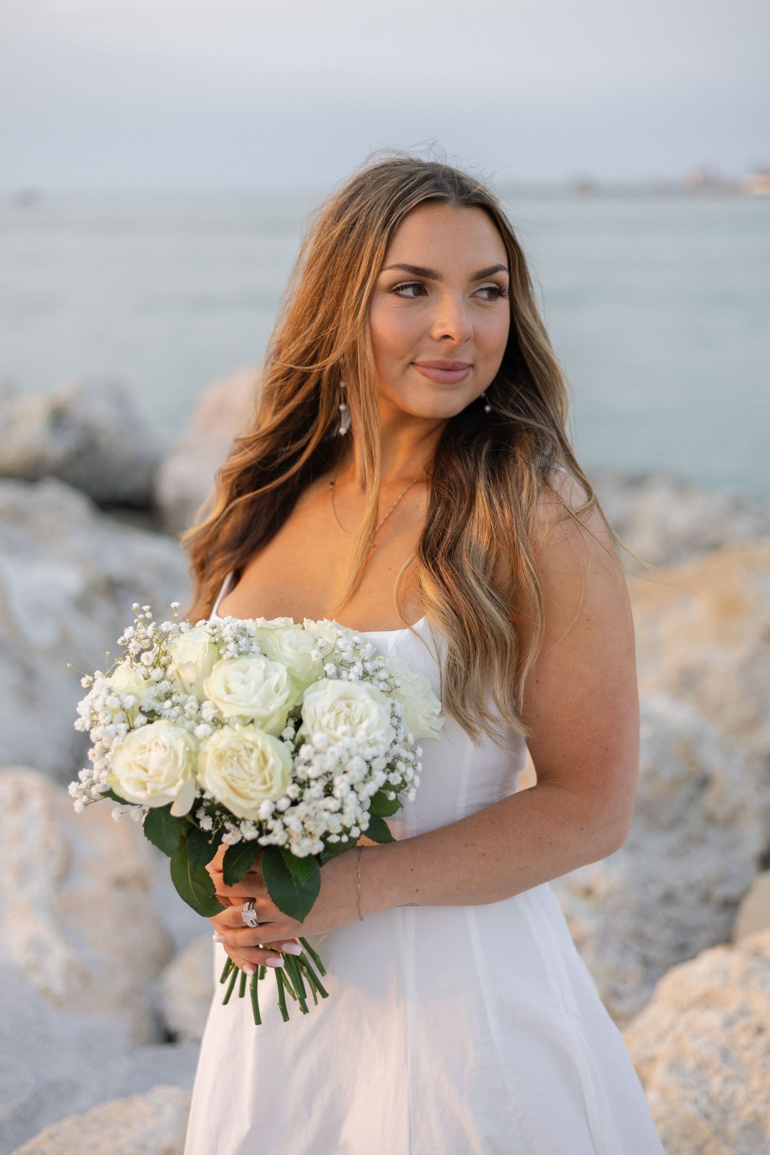 Bride in white strapless gown holds white rose bouquet by rocky beach at sunset.