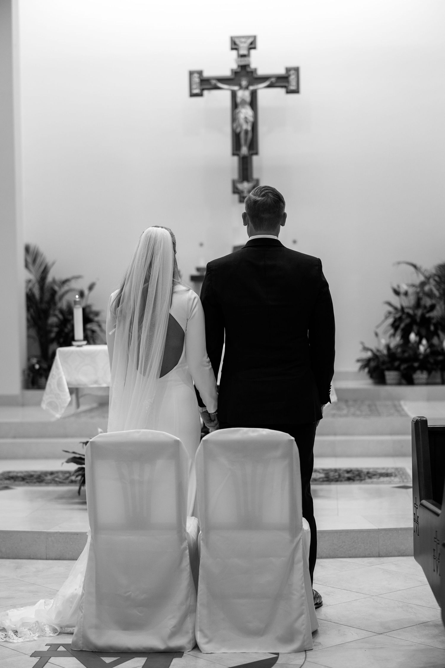 Black and white photo of bride and groom standing before crucifix at church altar.