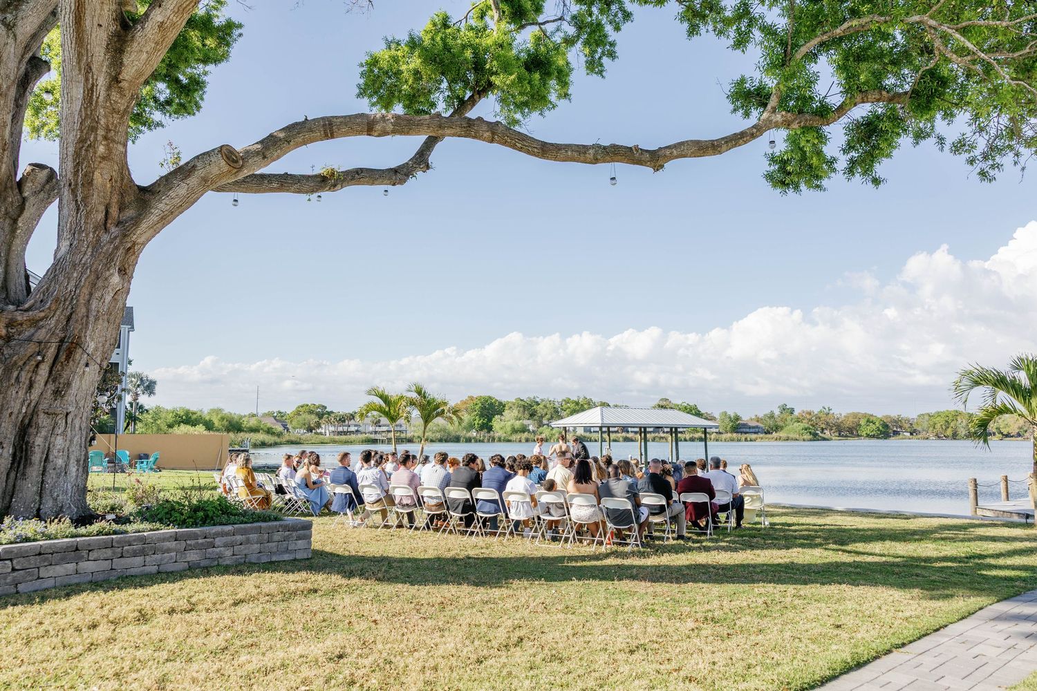 Outdoor waterfront wedding ceremony with guests seated under a large tree by a serene lake on a sunny day.