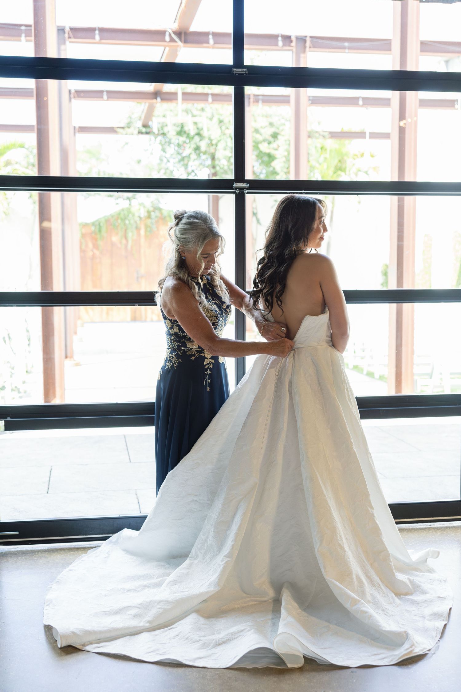 Mother helps bride button her elegant strapless wedding gown with a long train in front of large windows.