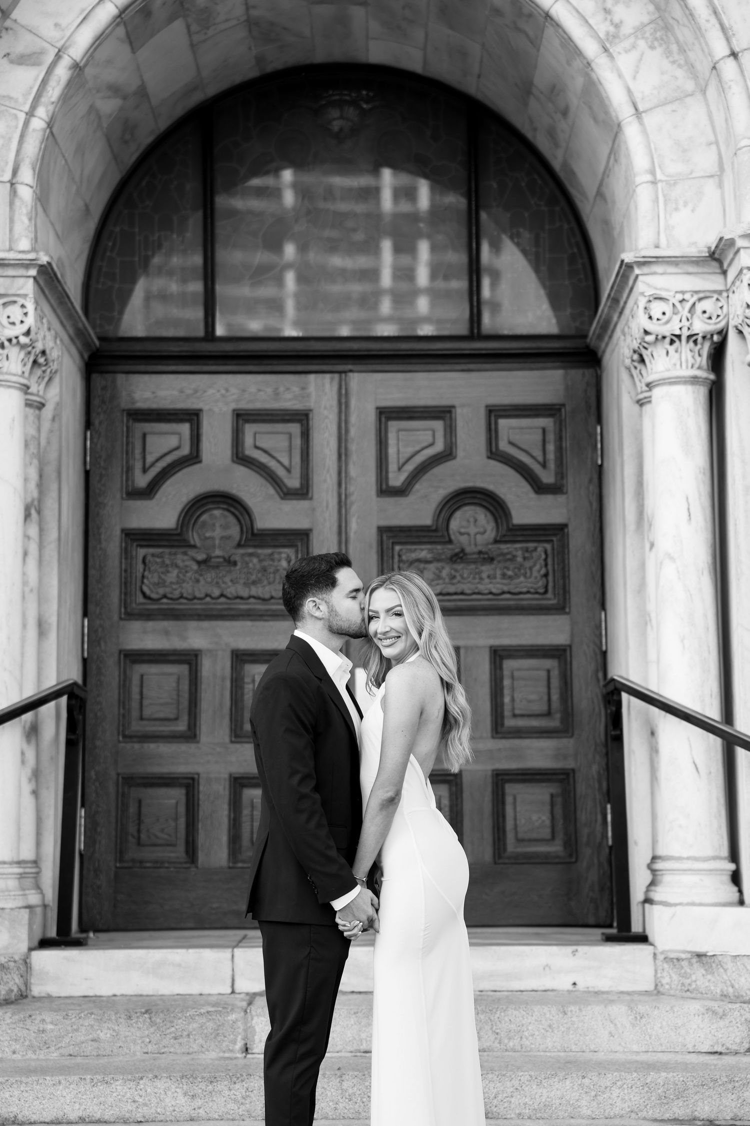 Black and white photo of bride and groom holding hands before ornate double doors of historic venue.