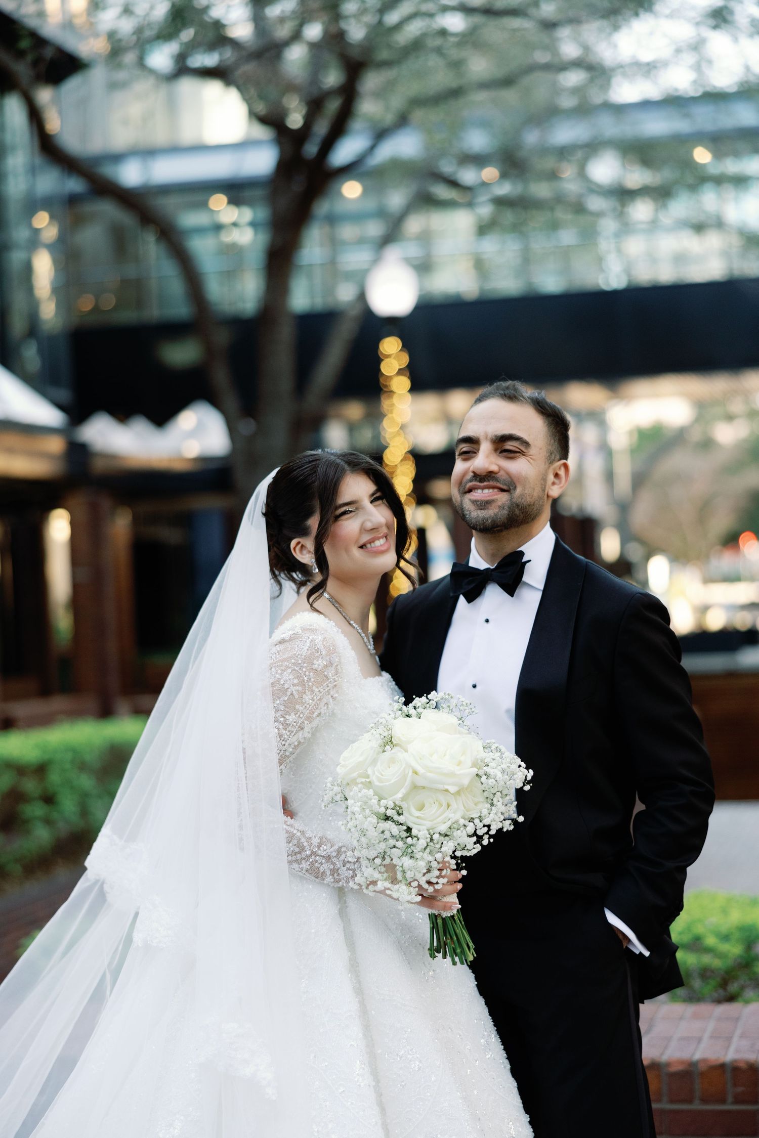 Smiling bride and groom posing outdoors with white bouquet in urban garden setting.