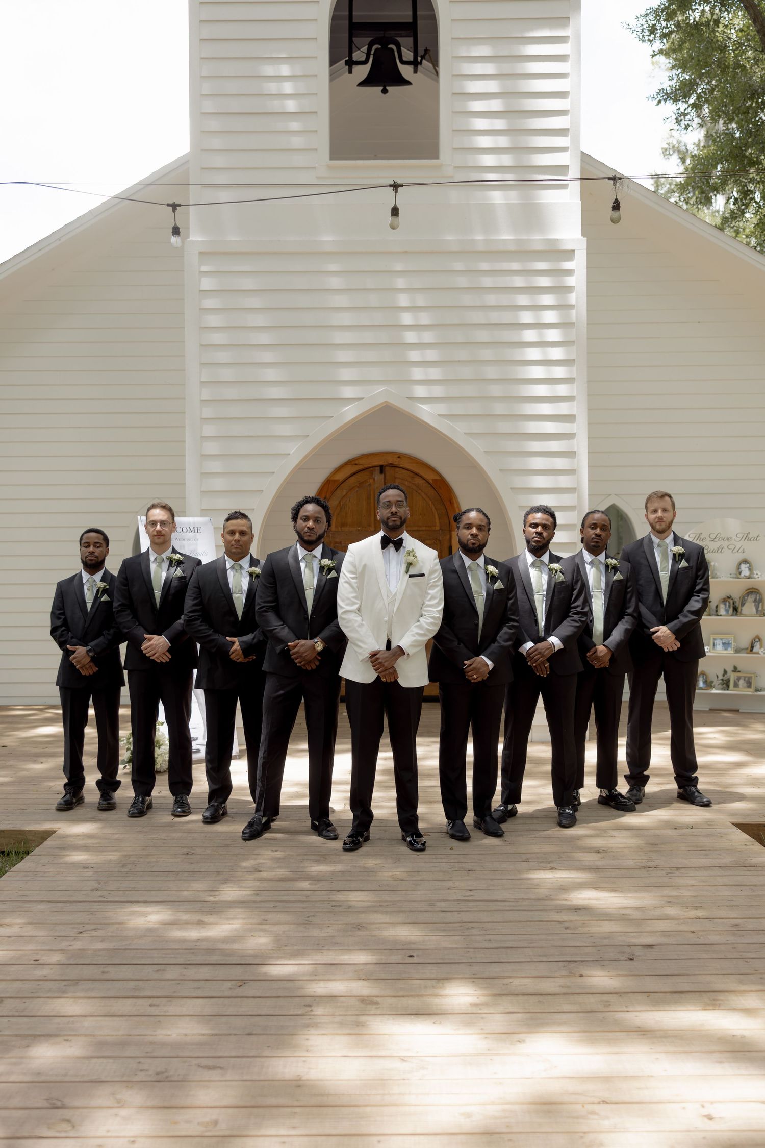 Groom in white jacket stands with ten groomsmen in black suits before white chapel with bell tower.