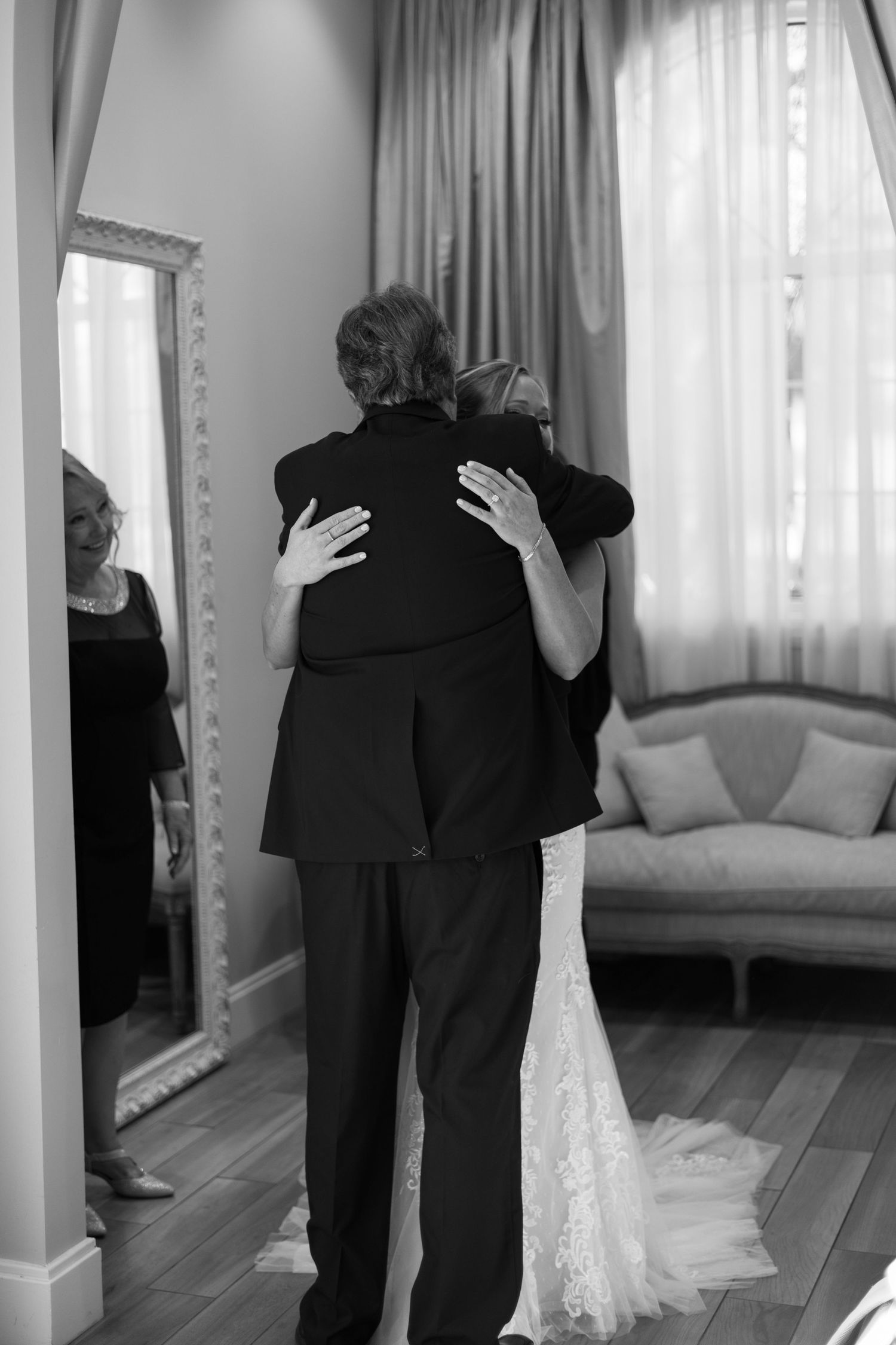 Bride in lace gown shares tender embrace with father in black suit beside full-length mirror.