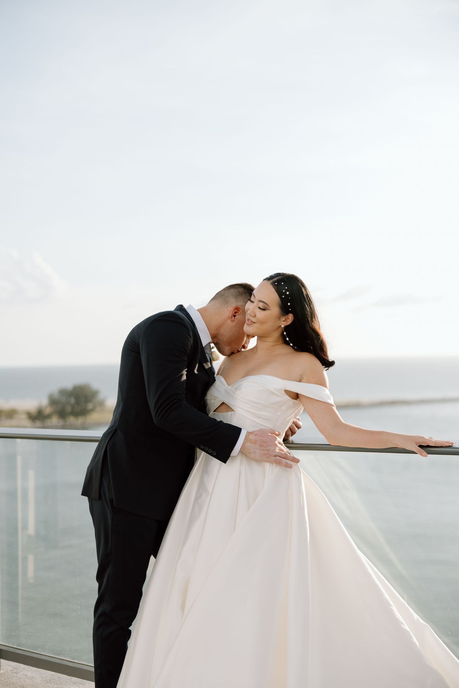 Groom in black suit kisses bride in off-shoulder white gown on waterfront balcony overlooking ocean.