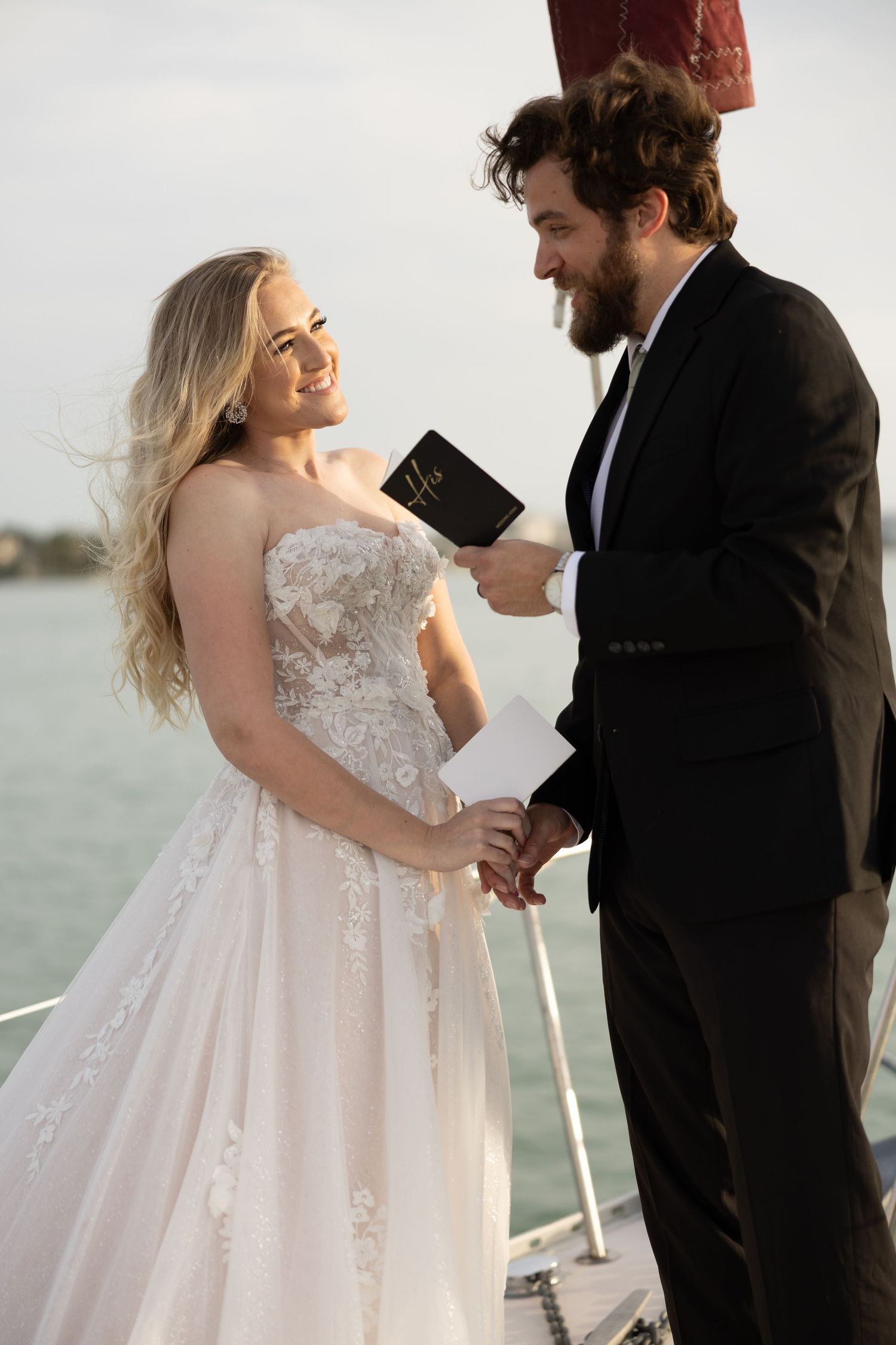 Smiling bride in lace gown holds hands with bearded groom reading vows on sailboat deck.