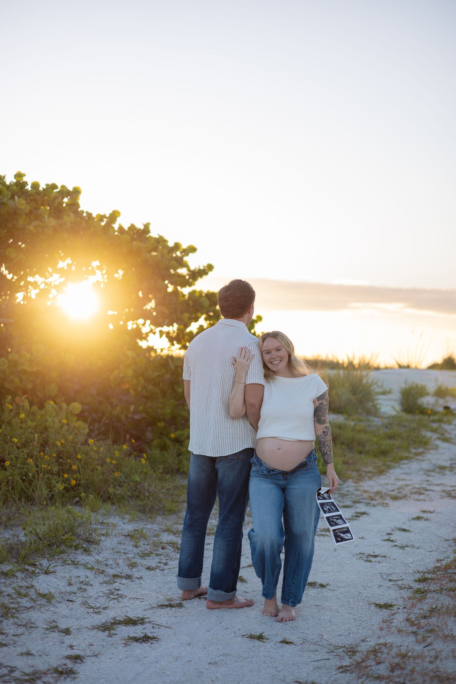 Expecting couple standing on sandy beach path at golden sunset, woman holding ultrasound photos.