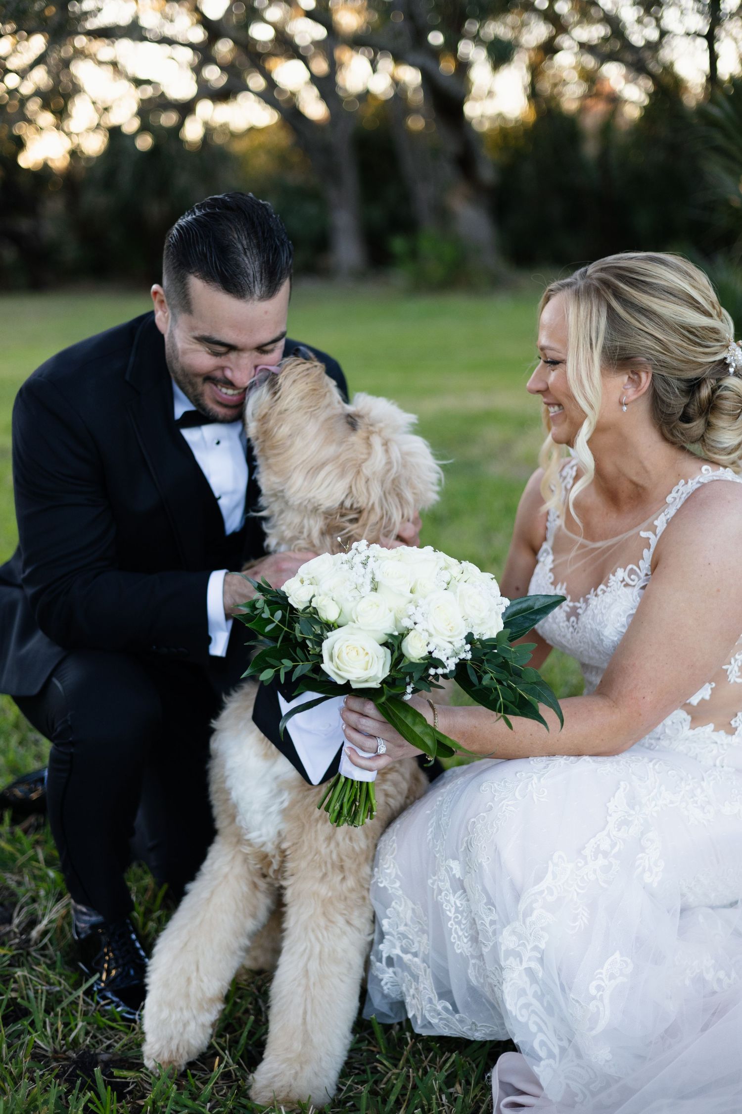 Smiling bride and groom kneel with their golden doodle dog wearing a bow tie at sunset.