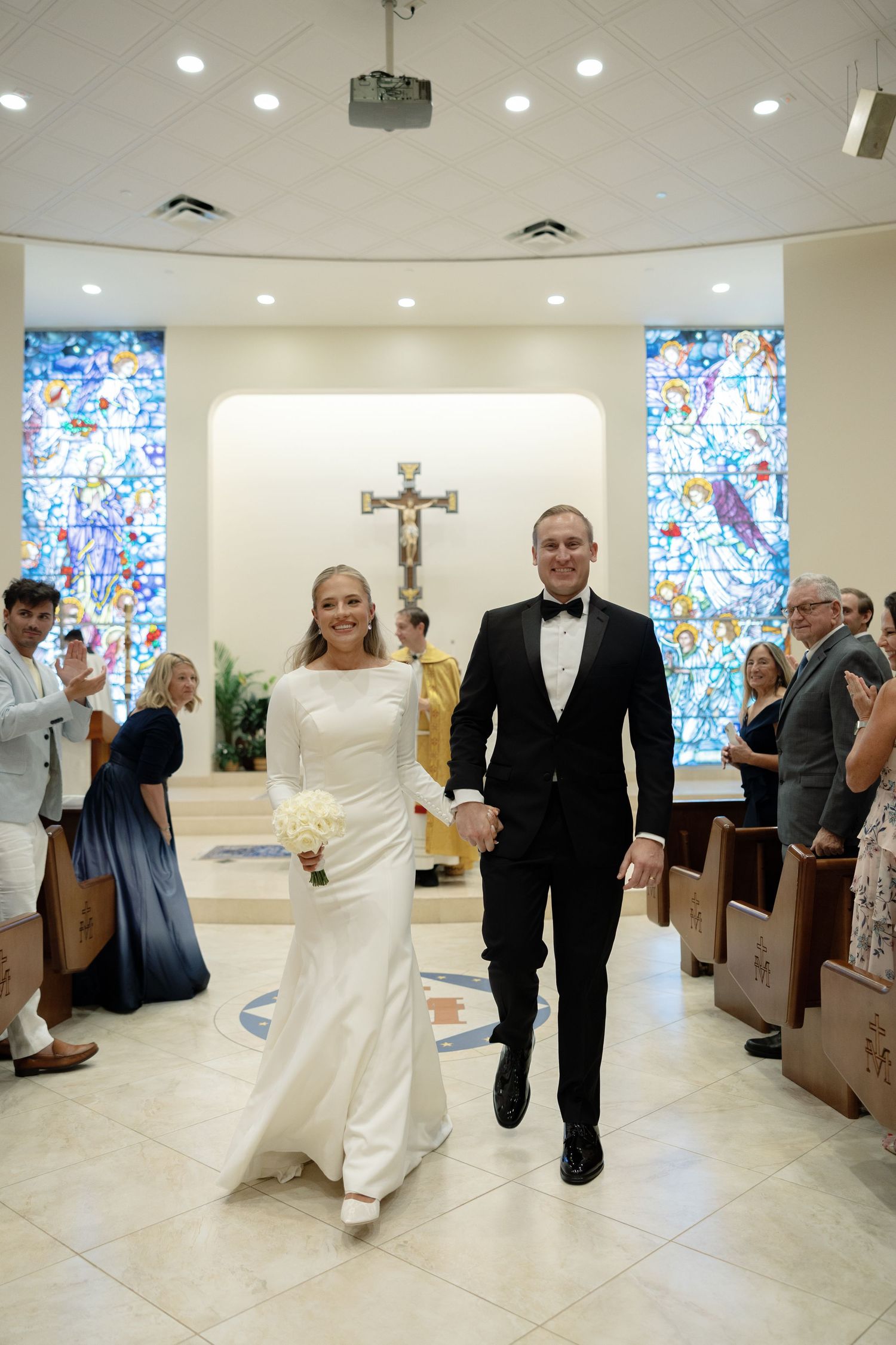 Newlyweds walk down aisle after wedding ceremony in modern church with colorful stained glass windows.