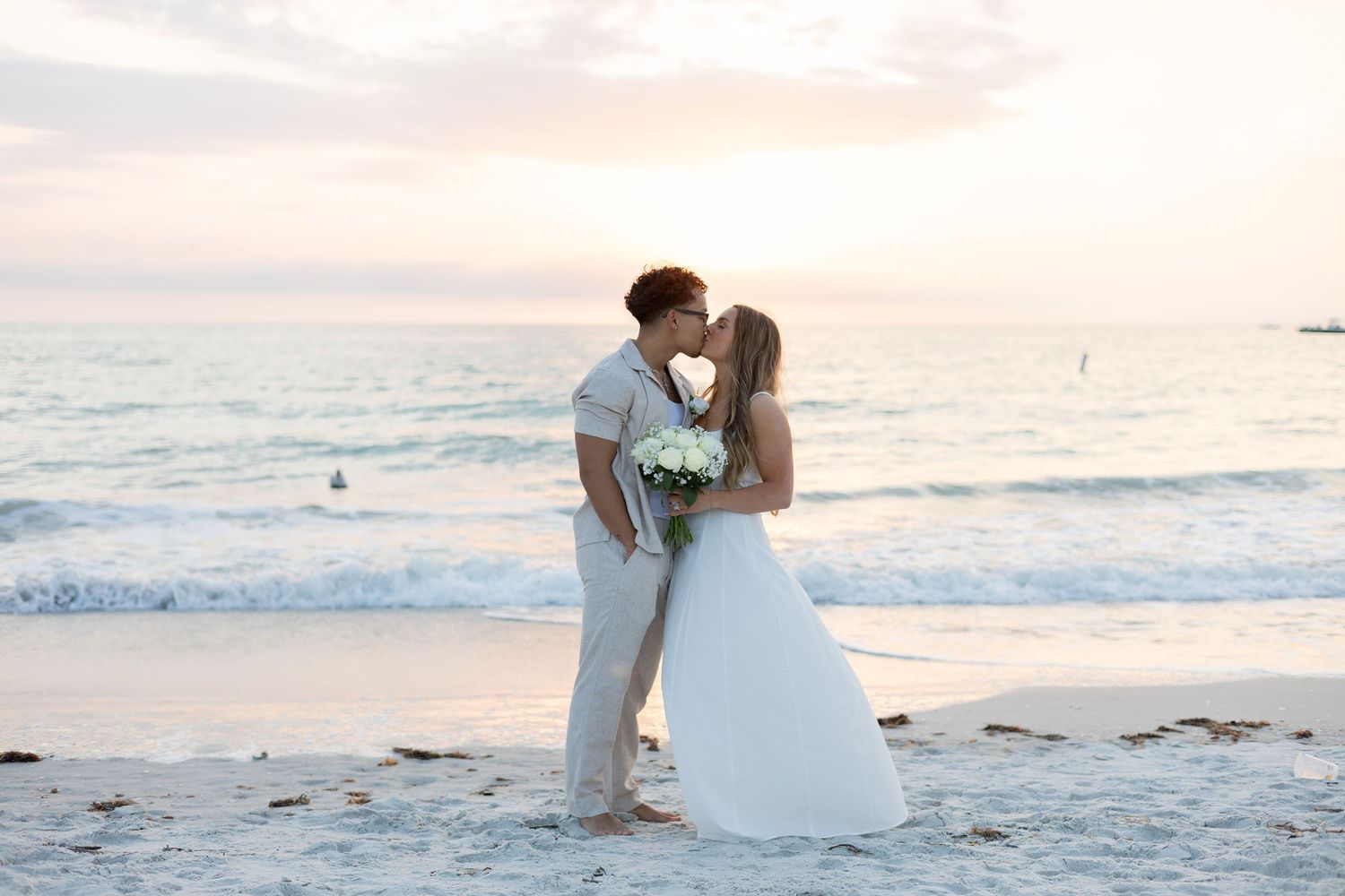 Newlyweds share first kiss on sandy beach with pastel sunset ocean backdrop.