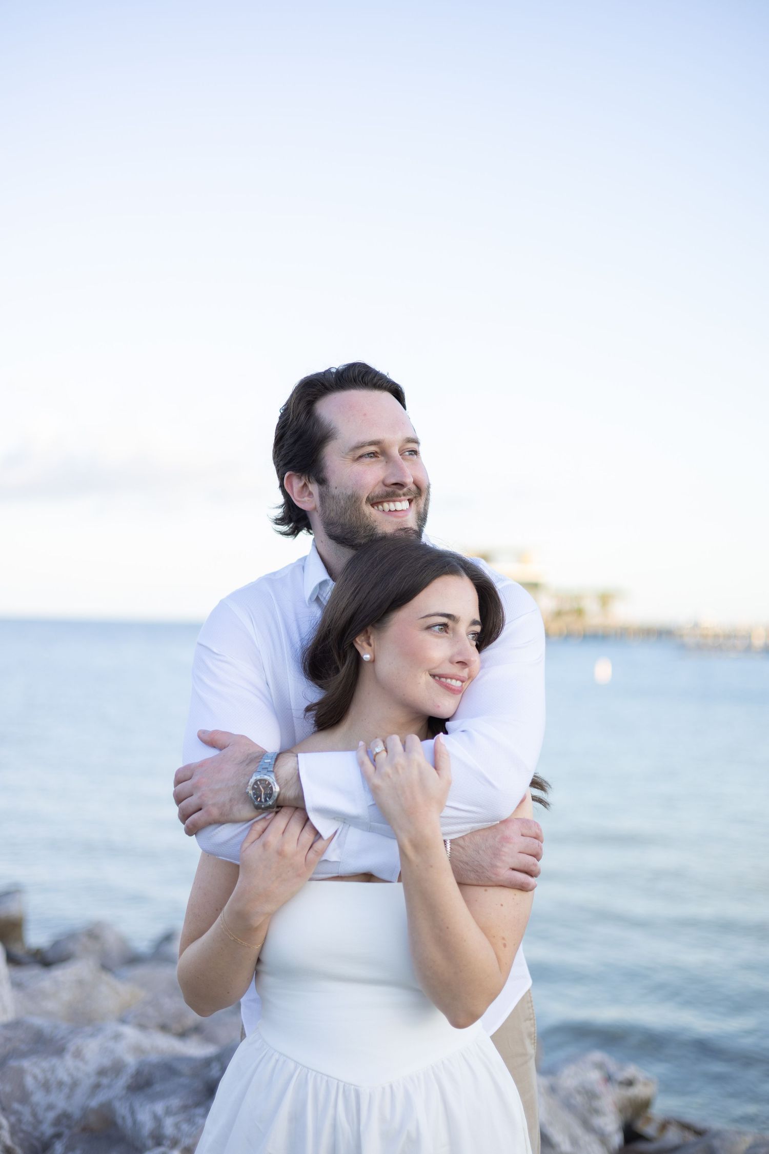 Happy couple embracing by the ocean at sunset, woman wearing white off-shoulder dress.