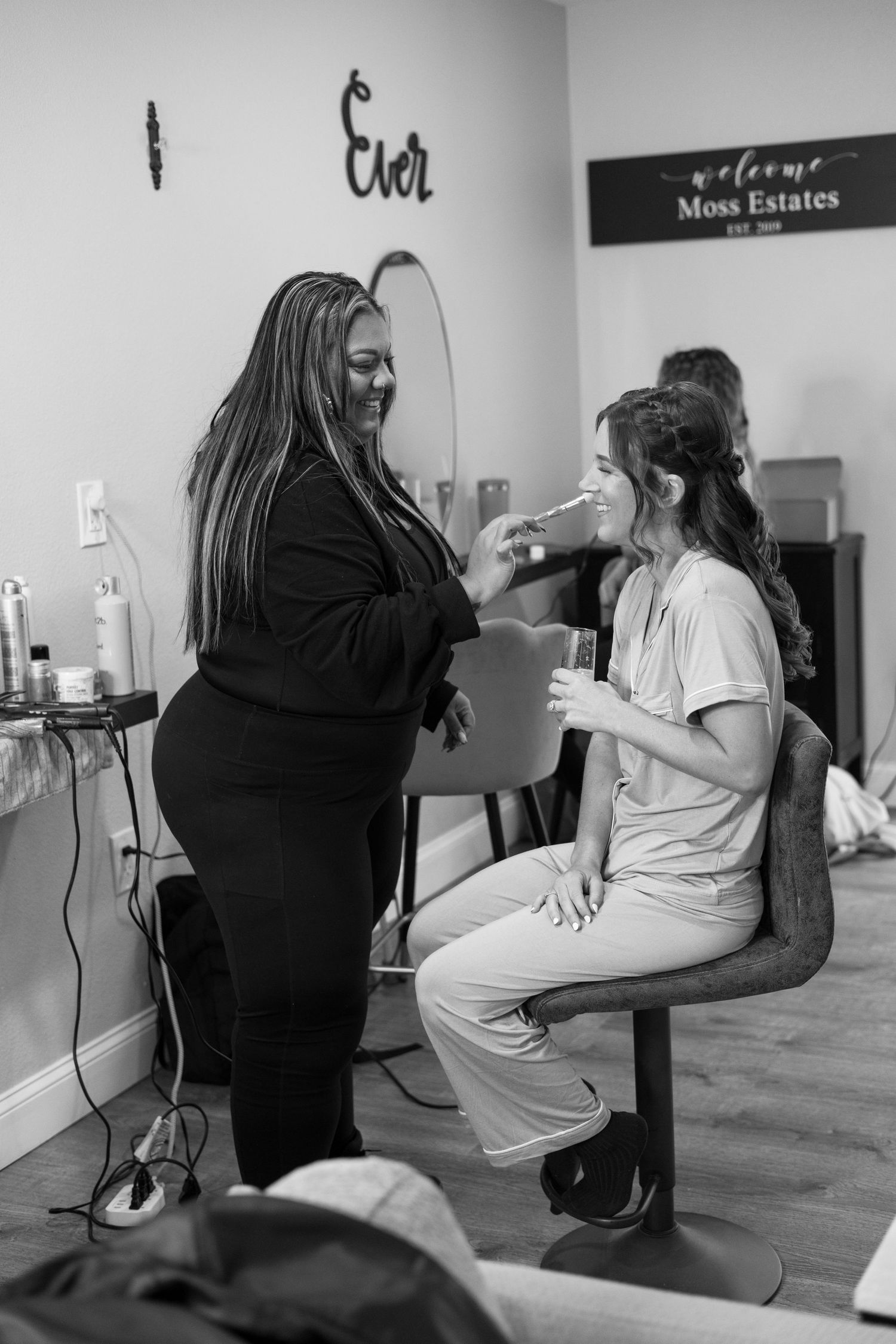 Black and white photo of makeup artist applying makeup to a smiling bride at Moss Estates salon.
