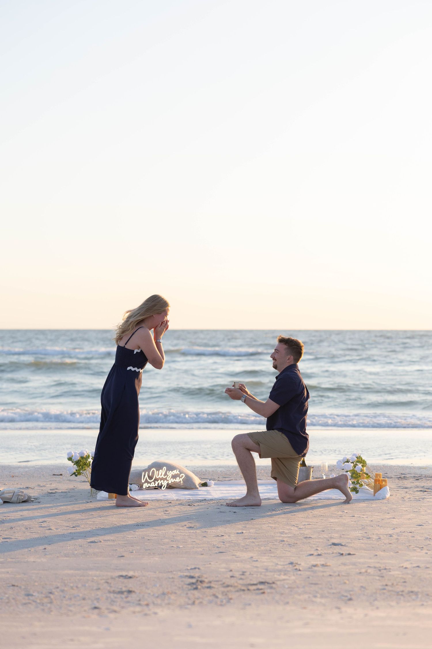Man proposes to woman on bended knee on a sandy beach at sunset with flowers nearby.