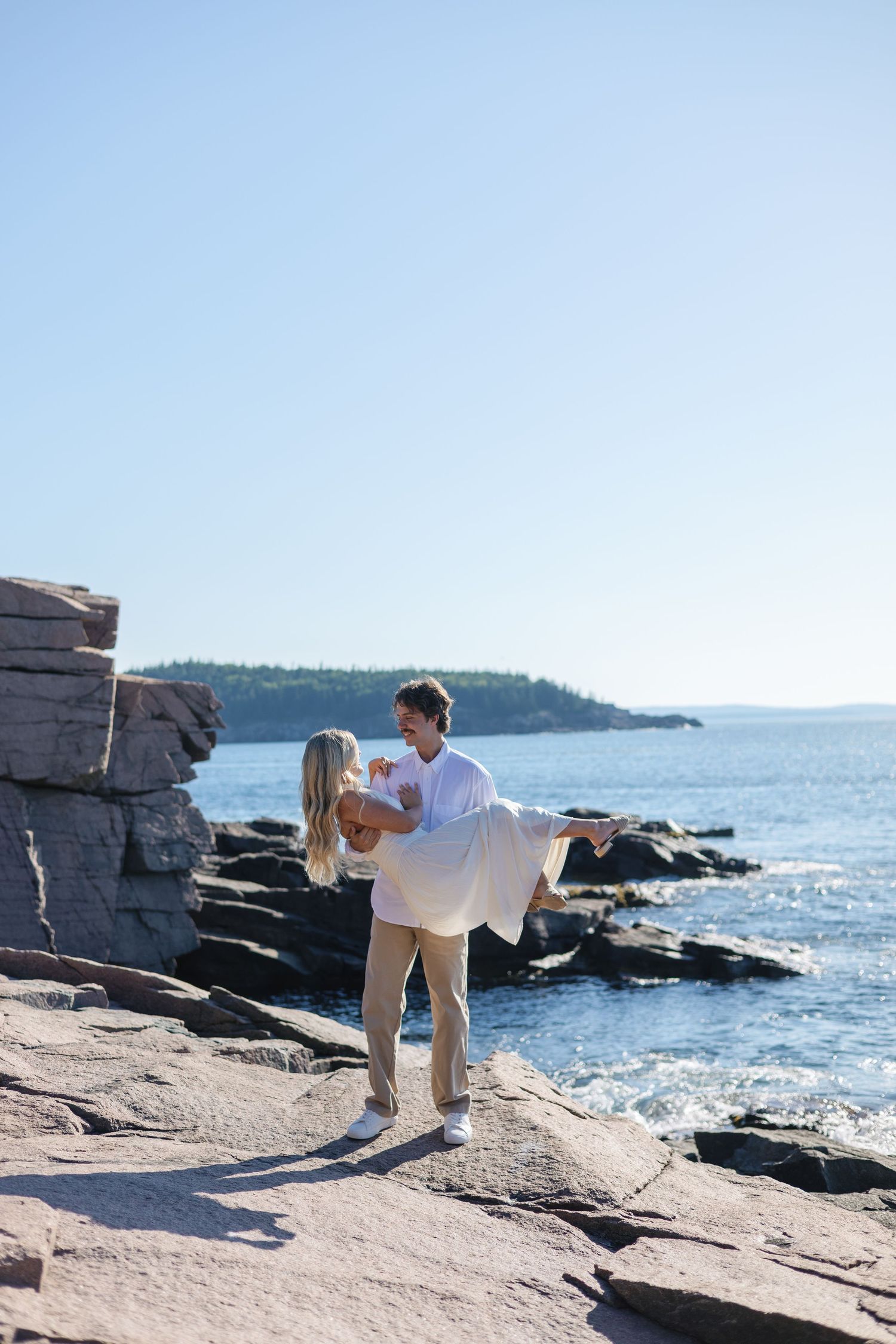 Man lifting woman in white dress on rocky ocean shore with island visible across calm blue water.