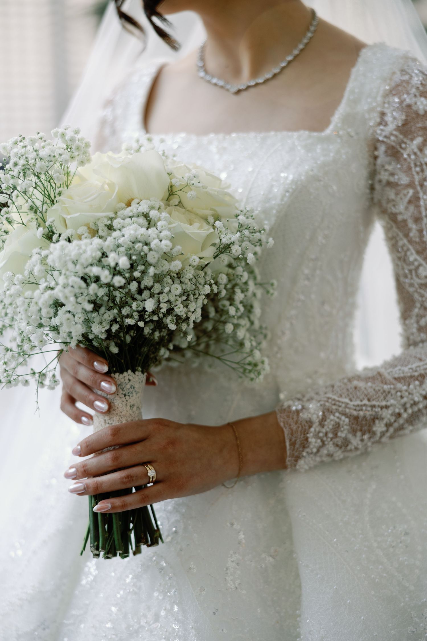 Bride in beaded lace gown holds white rose and baby's breath bouquet with gold wedding ring.