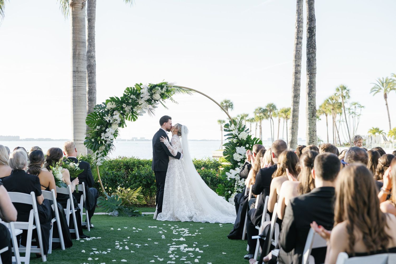 Couple kissing at outdoor wedding ceremony under floral arch with palm trees and ocean view, guests seated in white chairs.