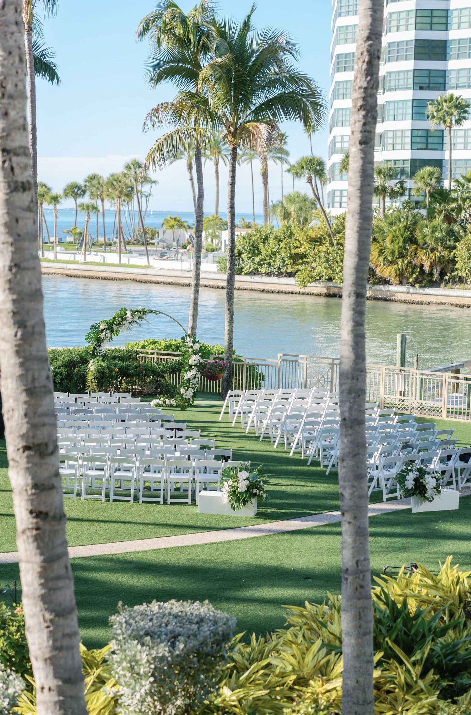 Outdoor waterfront wedding ceremony setup with white chairs, floral arch, palm trees, and ocean views at a tropical venue.