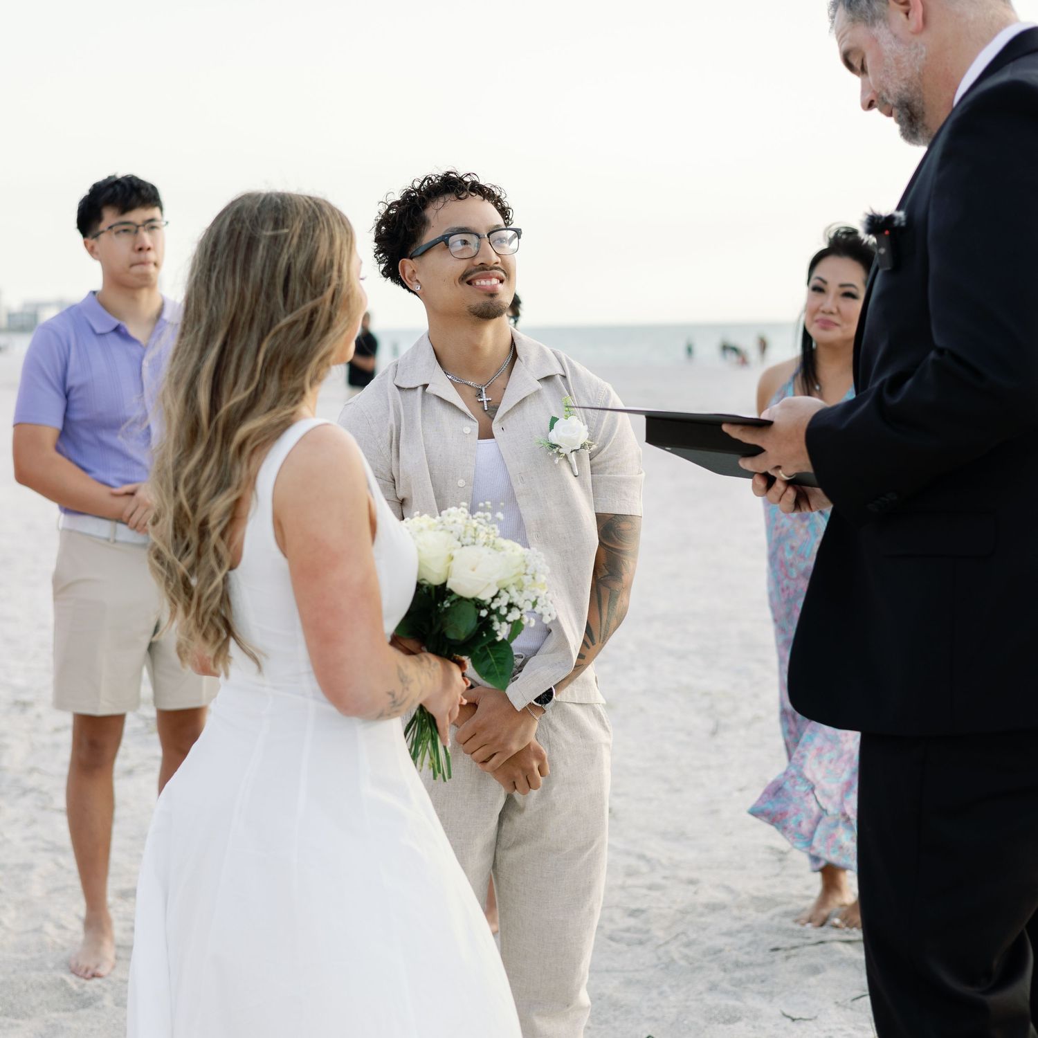 Groom smiles at bride during intimate beach wedding ceremony with small group.